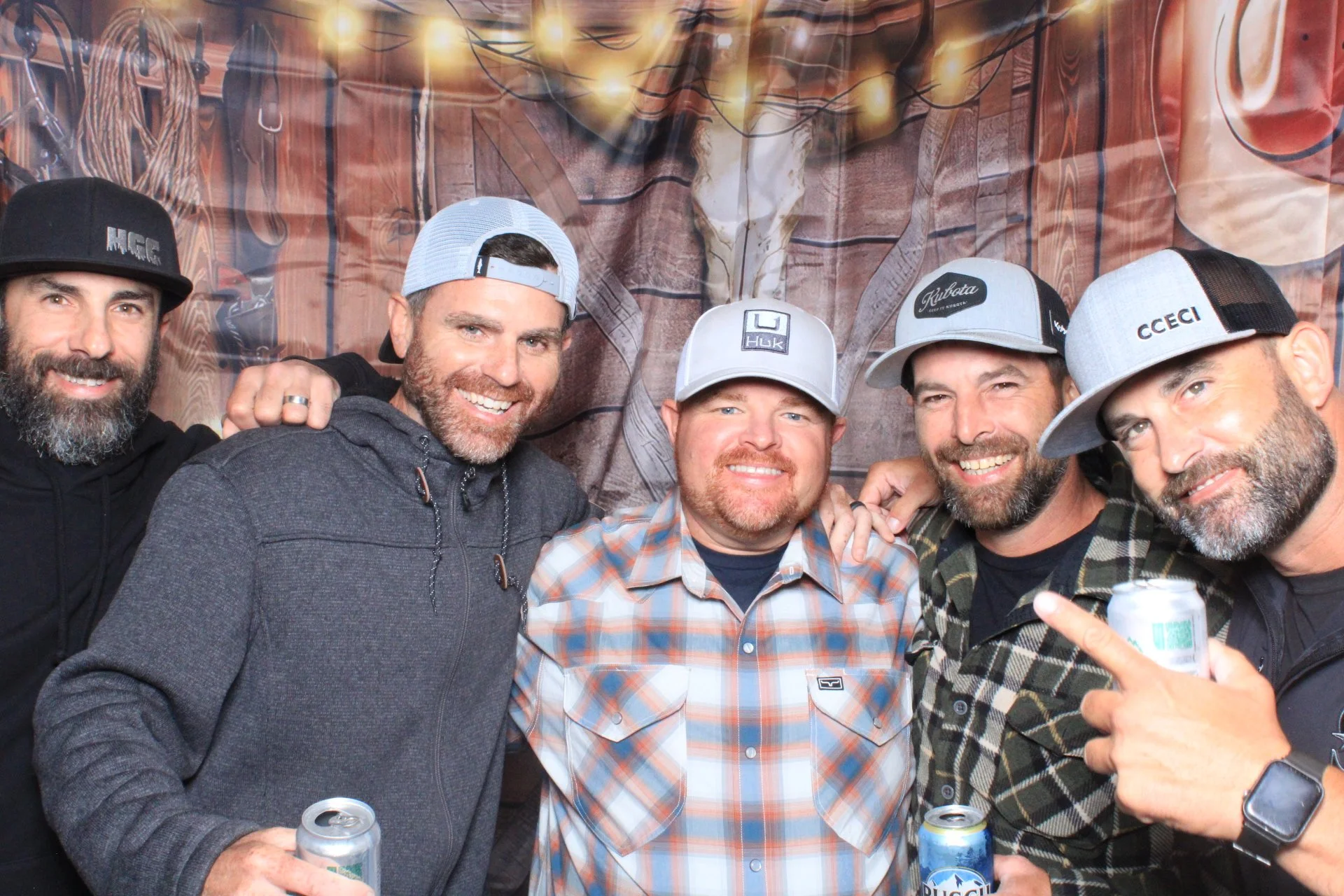 Six men posing together indoors with a rustic wooden backdrop and string lights, all wearing casual winter clothing and baseball caps. Some are holding cans of beer, smiling and having a good time.