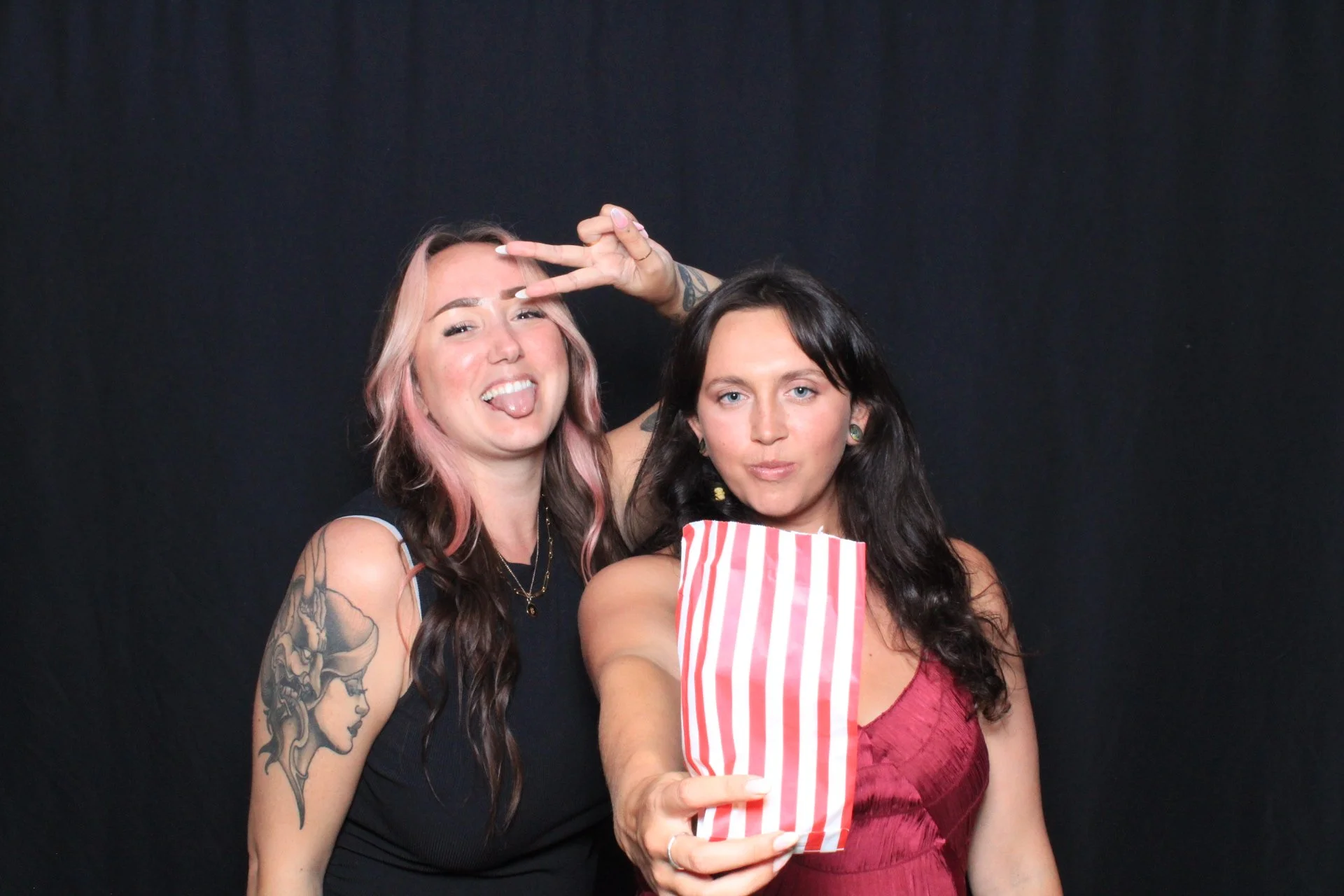 Two young women posing for a photo against a black background. The woman on the left has long, wavy hair with pink highlights, visible tattoos on her arm, and is sticking out her tongue while making a peace sign with her hand. The woman on the right 