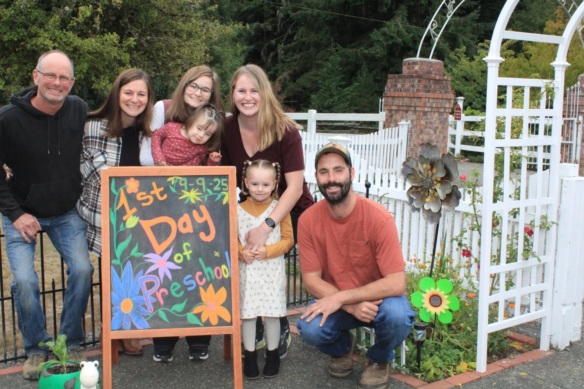 A group of people celebrating the first day of preschool, with a colorful sign and decorative flowers in a garden setting.