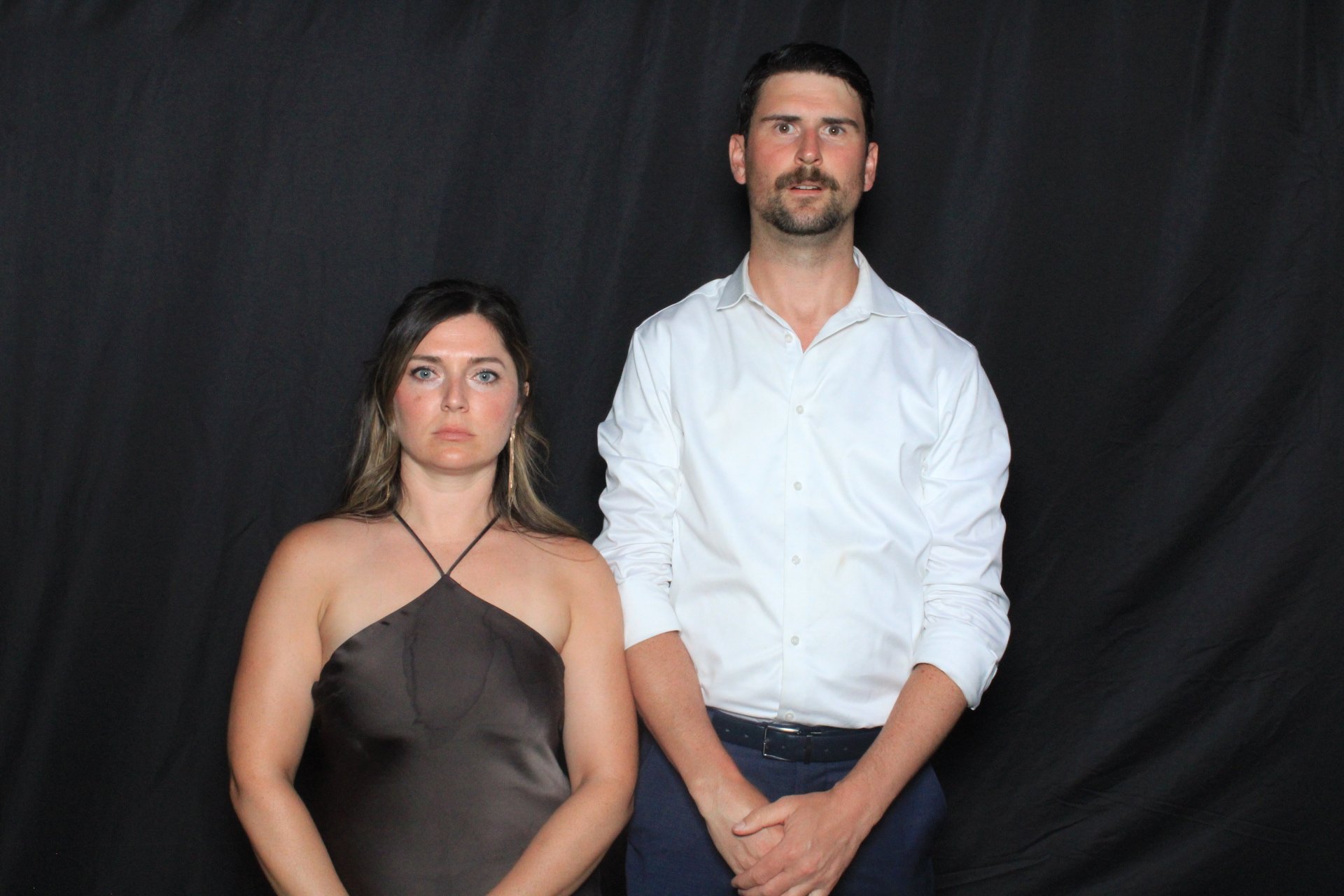 A woman in a black satin halter dress and a man in a white dress shirt standing against a black background, both looking at the camera with serious expressions.
