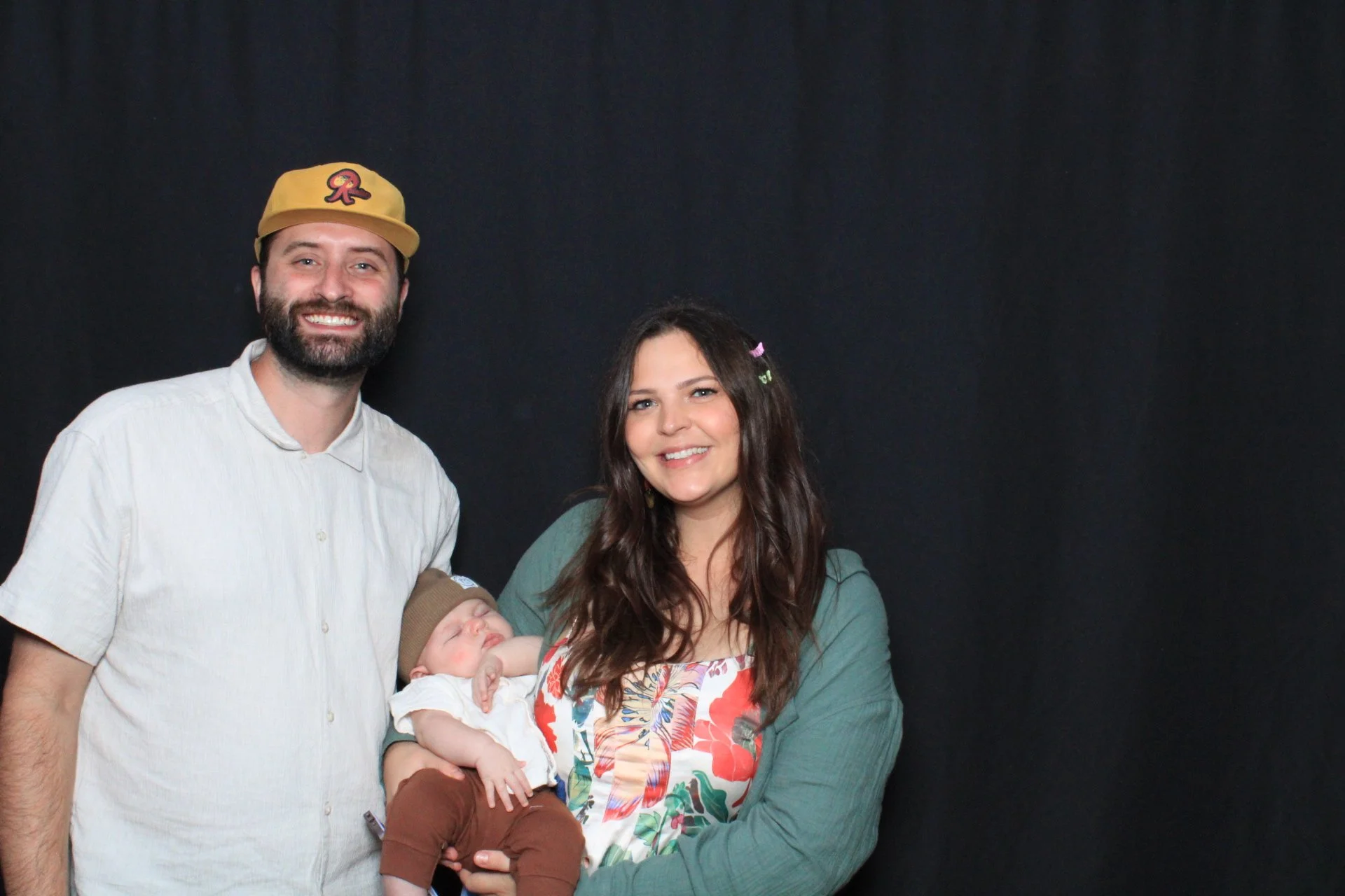 A family of three: a man with a beard and baseball cap, a woman with long brown hair, and a sleeping baby in her arms, standing against a black background.
