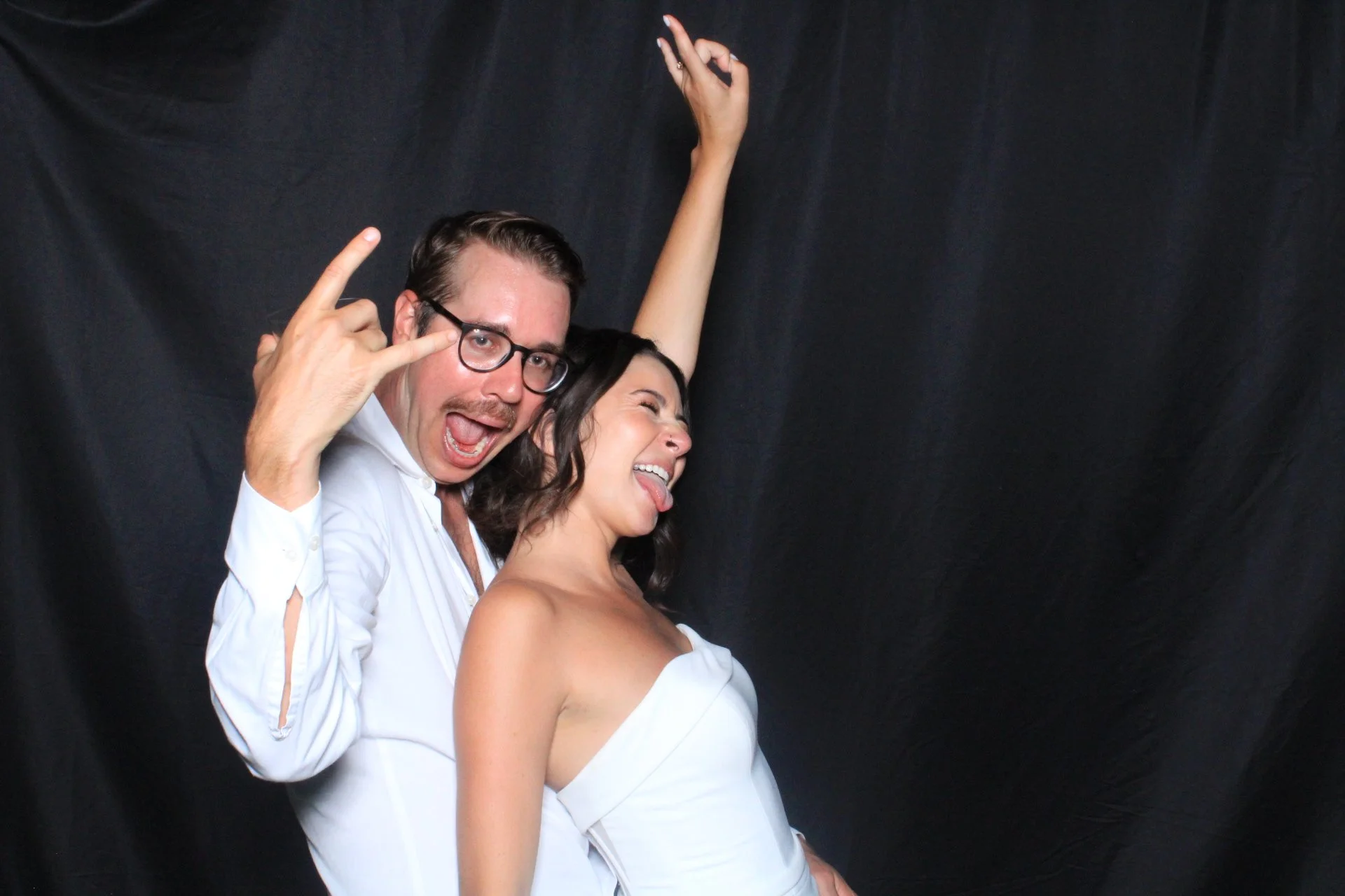 Two people, a man with glasses and a woman, are posing playfully against a black backdrop. The man is making a rock 'n' roll hand gesture and has his mouth open, while the woman is sticking out her tongue and leaning back with her eyes closed.