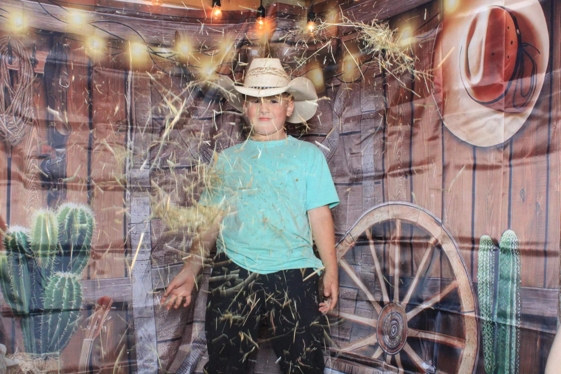 A boy standing inside a rustic barn with a large wagon wheel, cacti, and cowboy hats on the walls. He wears a cowboy hat and a light blue shirt, and there are sparklers burning around him.