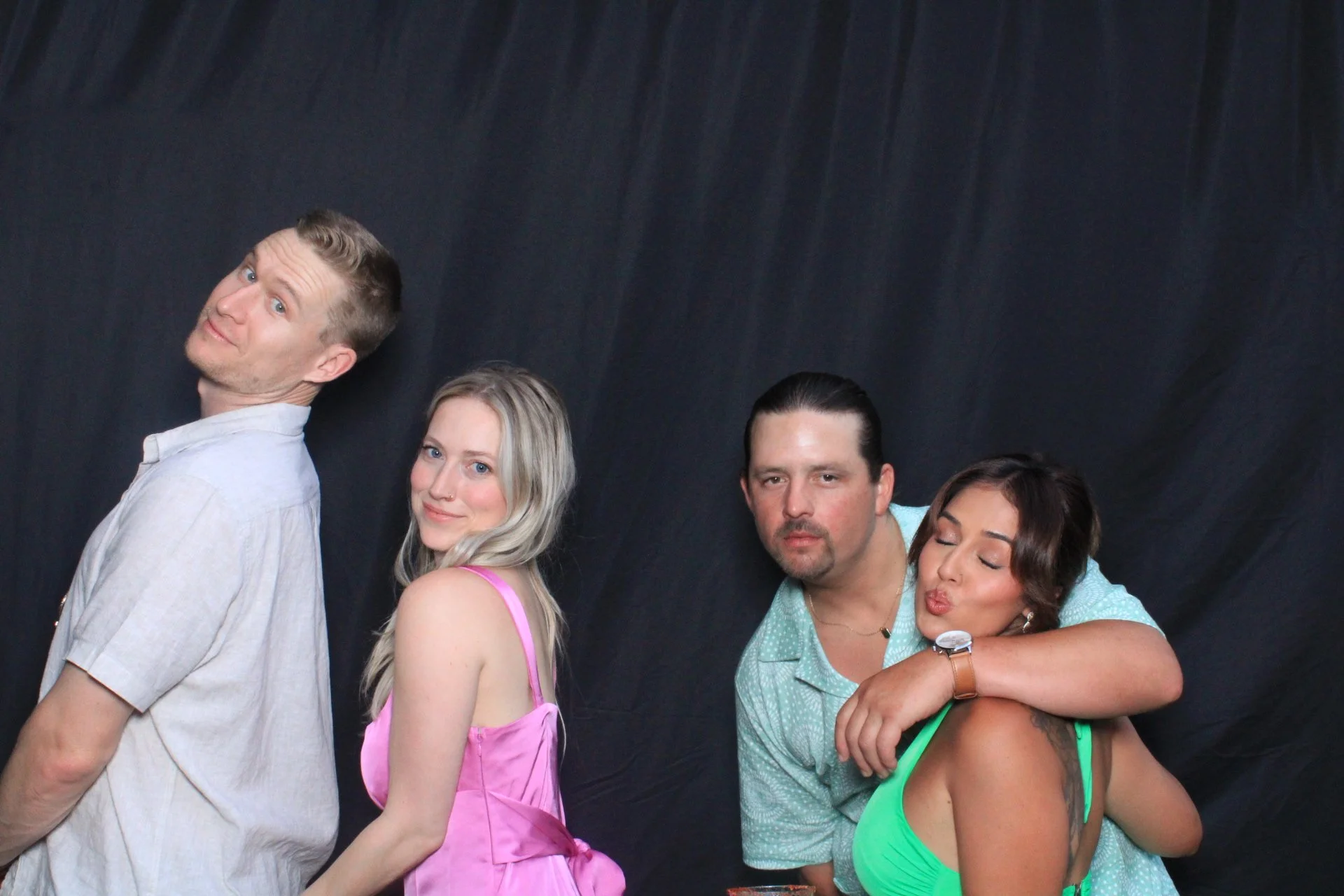Four friends standing in a line against a dark background, posing for a photo. The first person is a man with blonde hair, wearing a light-colored shirt, smiling and looking over his shoulder. The second person is a blonde woman in a pink dress, smil