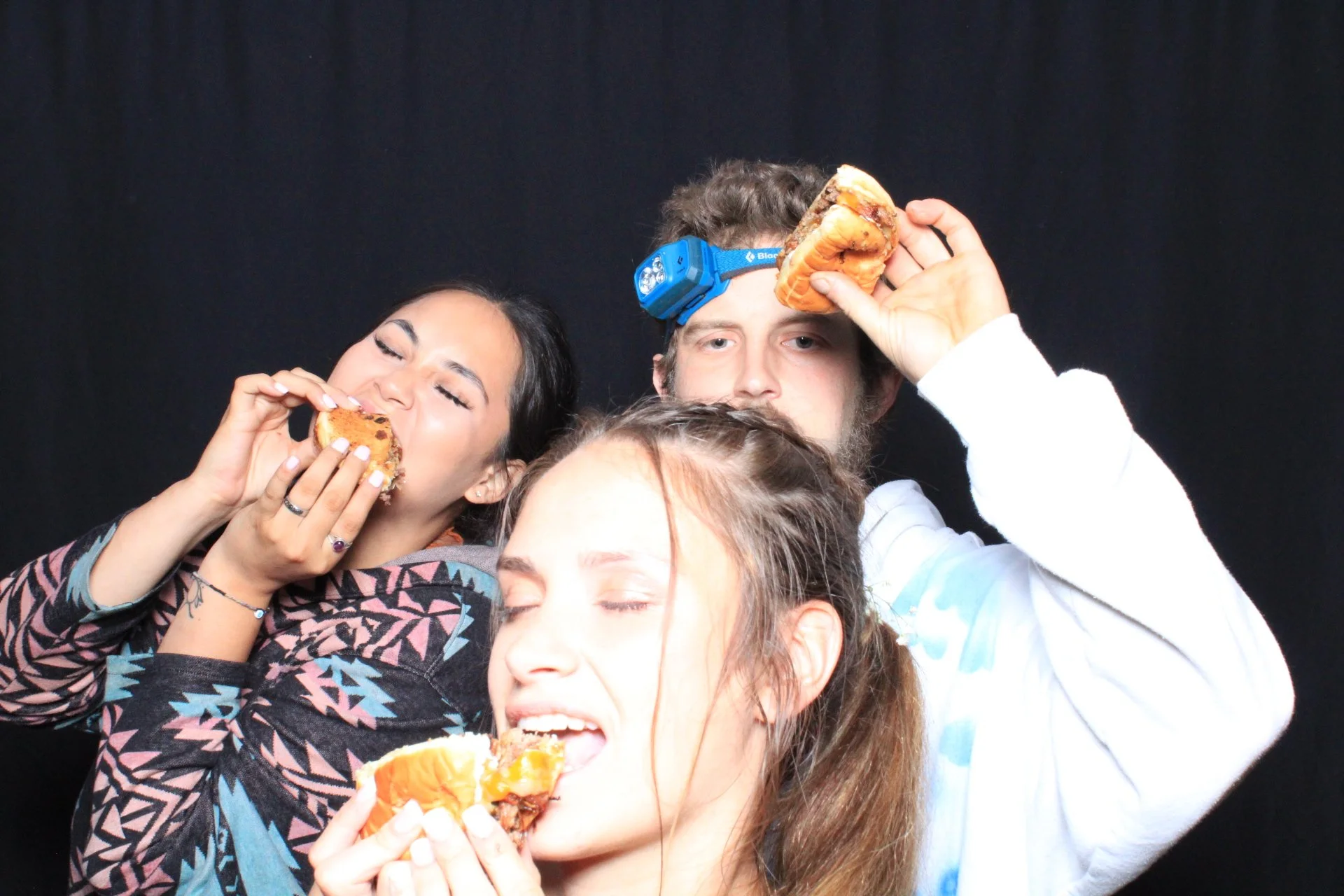 Three young adults eating pizza against a black background, with one person wearing a headlamp.