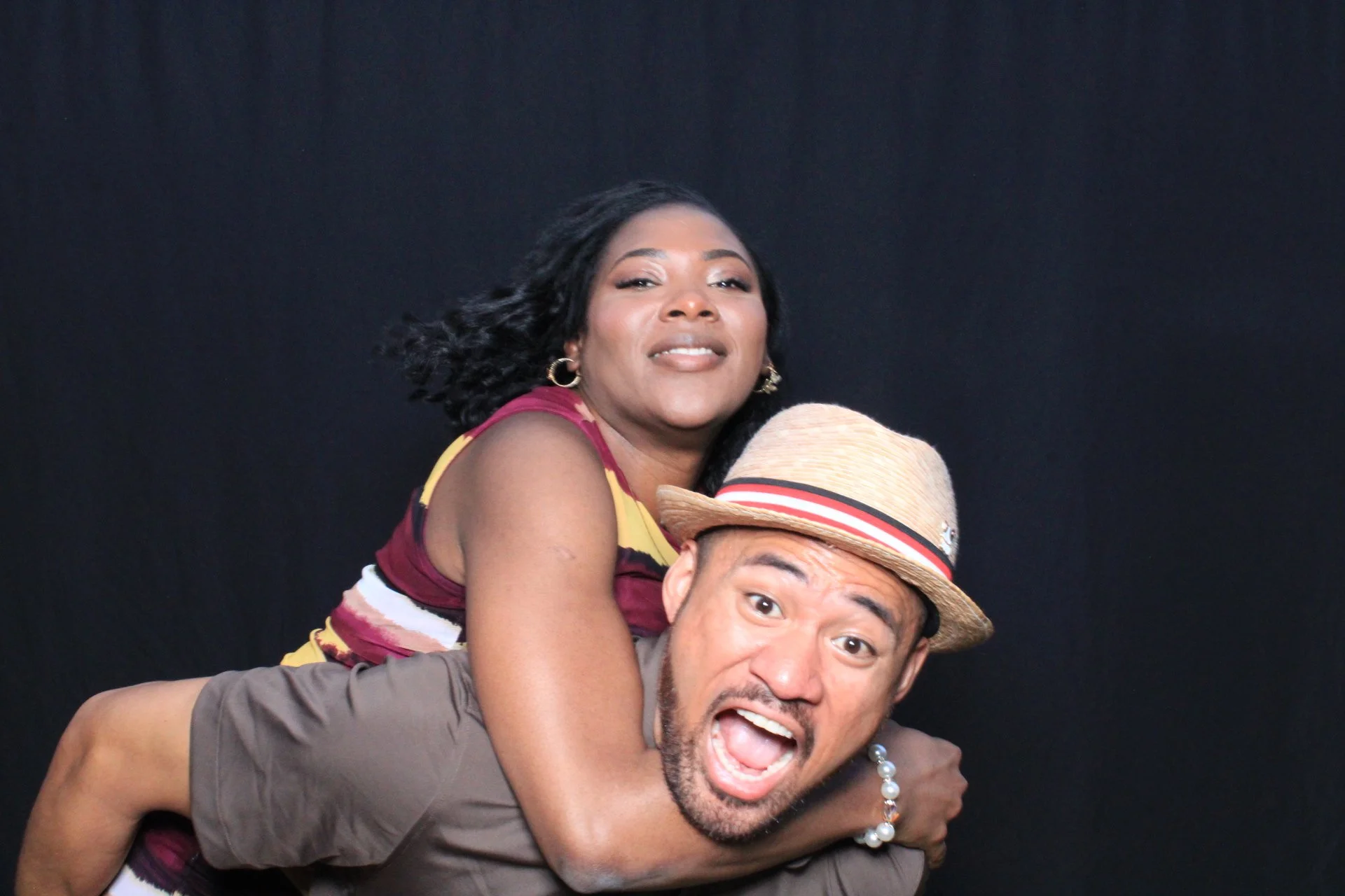 A woman with dark hair and hoop earrings sitting on a man's shoulders. The man is wearing a straw hat and has a beard, with his mouth open and eyes wide. The background is black.