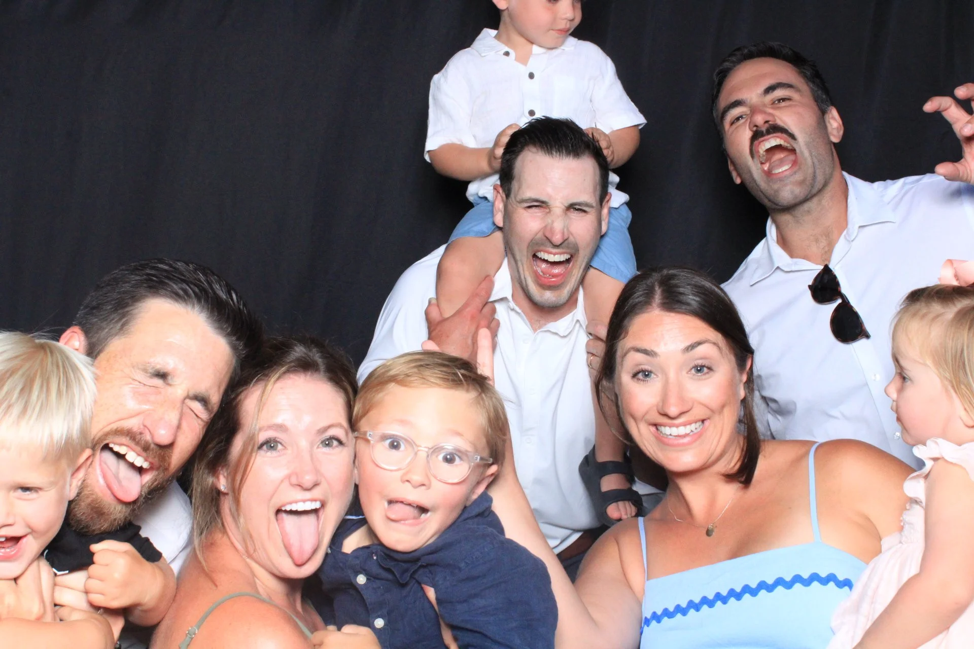 A group of smiling adults and children posing together, some making faces and sticking out their tongues, against a black background.