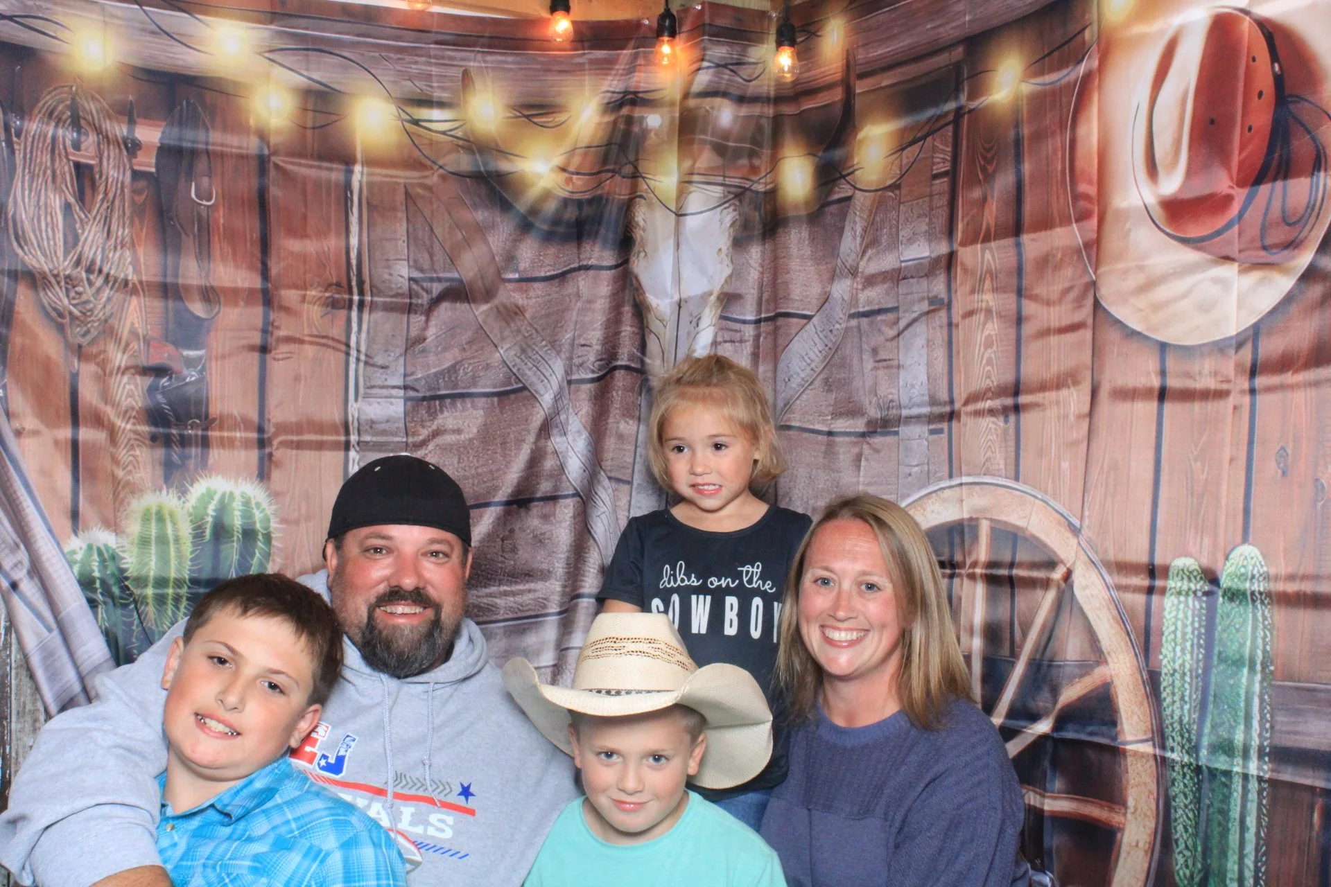 A family of five posing in front of a rustic cowboy-themed backdrop with string lights, cacti, wagon wheel, and cowboy hat, at a cowboy-themed event.