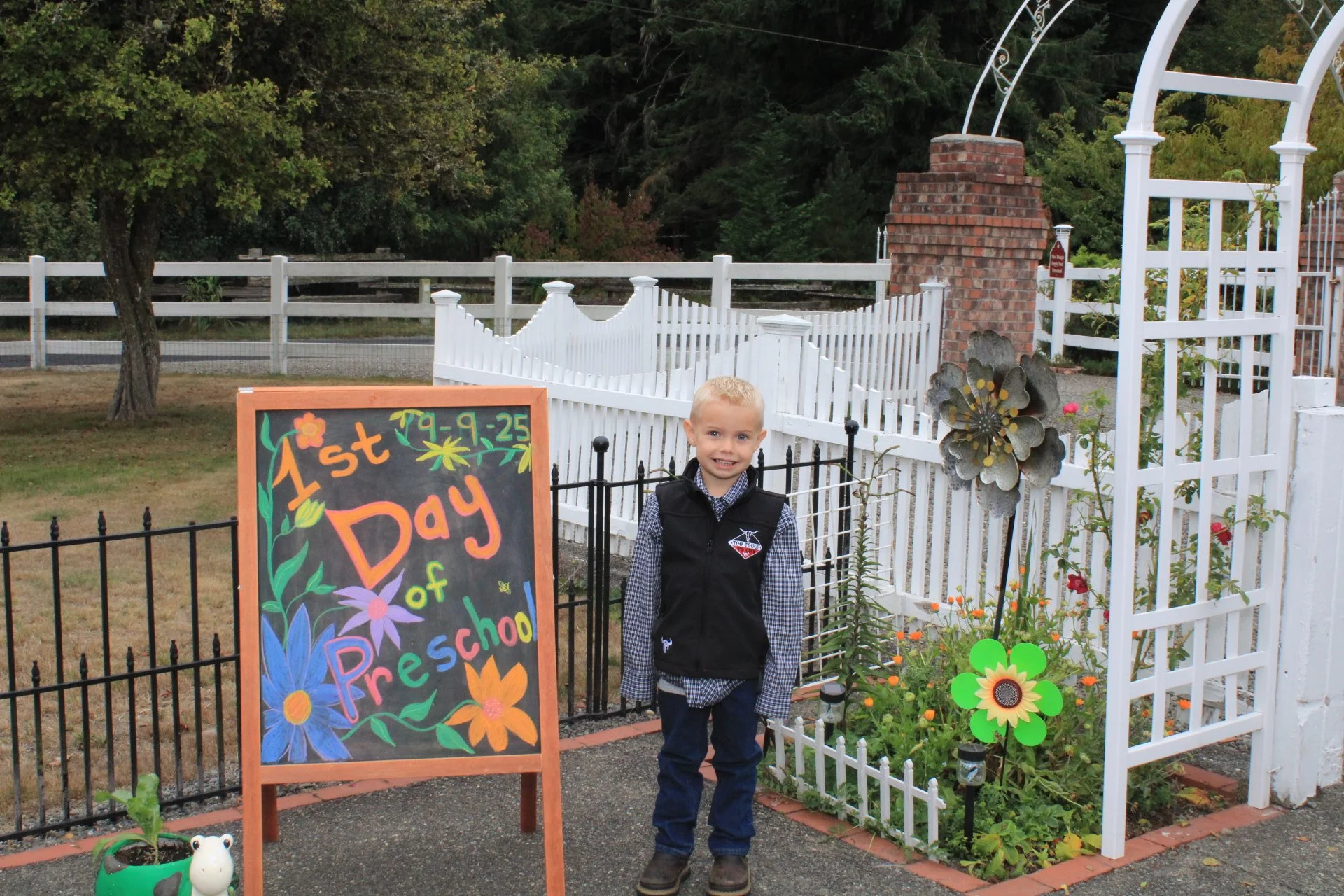 A young boy standing in front of a white fence and garden, smiling, next to a sign that reads '1st Day of Preschool 79-9-25'.