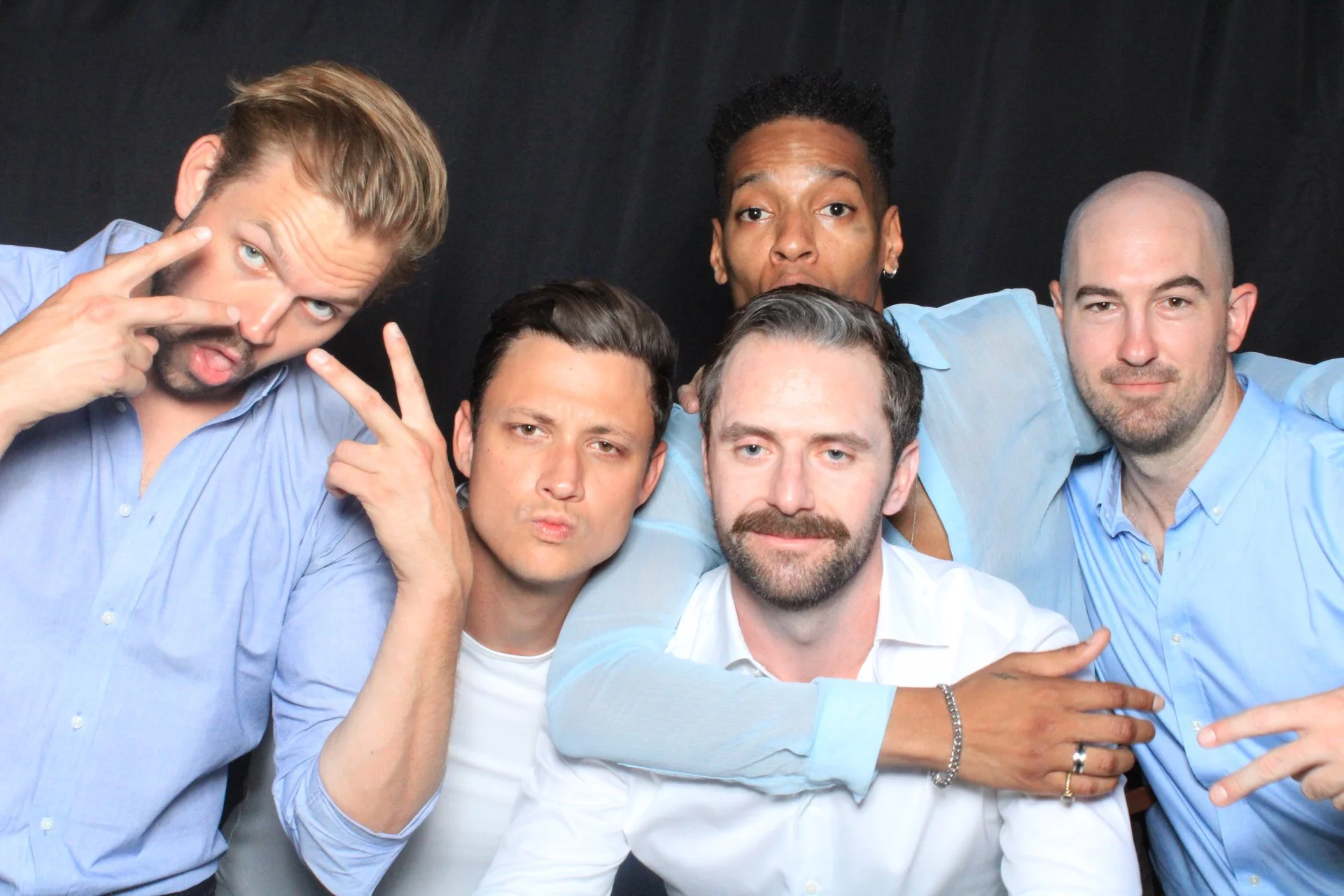 Group of five men with light and dark skin tones wearing casual shirts, posing and making expressive gestures against a black background.