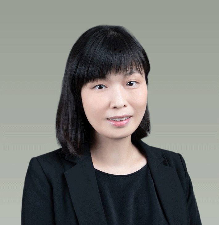 A young woman with black hair and wearing a black blazer and top, smiling softly against a light gray background.