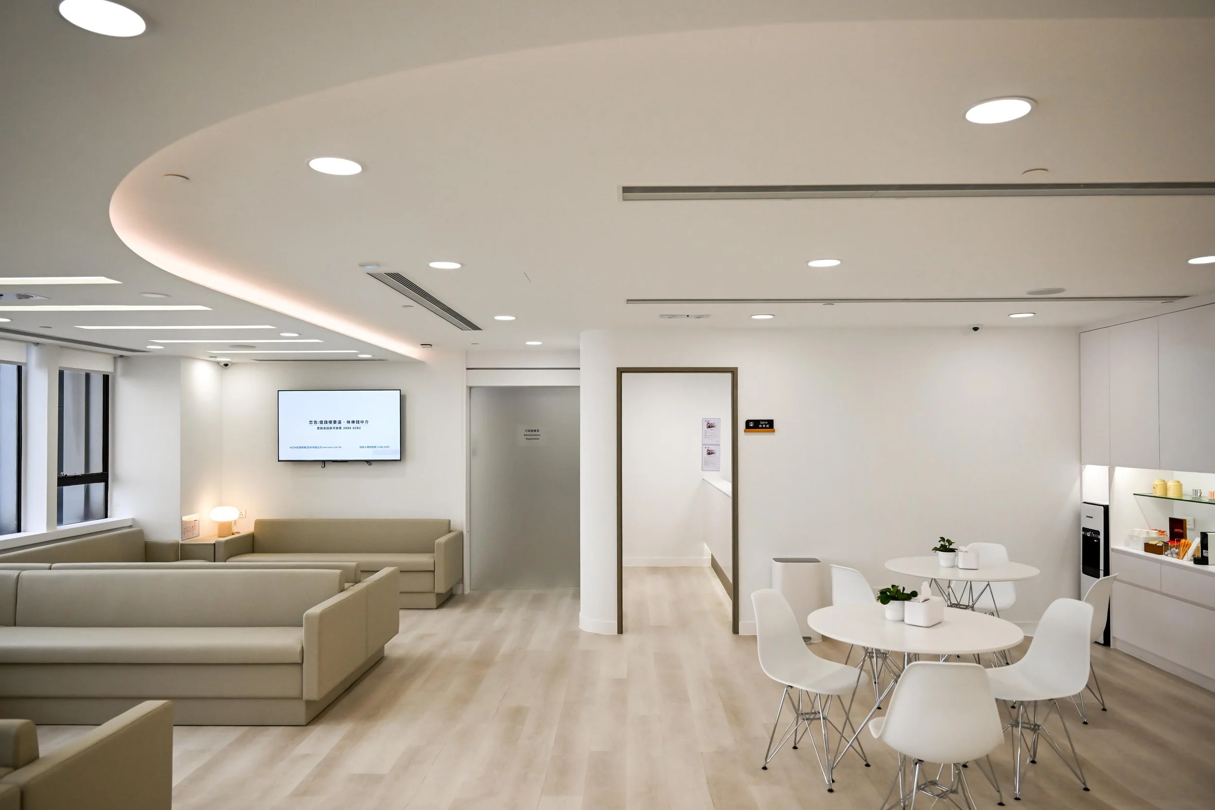 Modern, minimalistic lounge area with white walls, beige sofas, round white tables with white chairs, a wall-mounted TV, and a small kitchenette with a coffee machine and shelves with cups and containers.