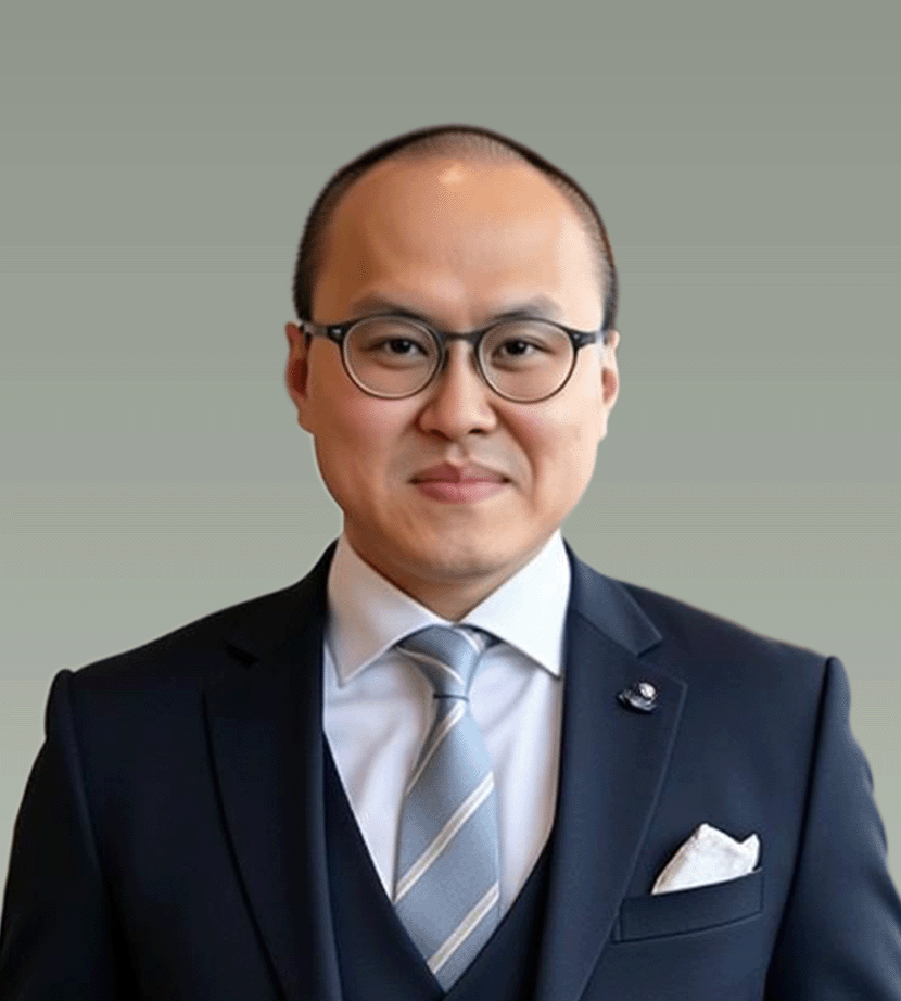 A professional man wearing glasses, a dark suit with a light-colored shirt, a striped tie, and a pocket square, posing against a plain gray background.