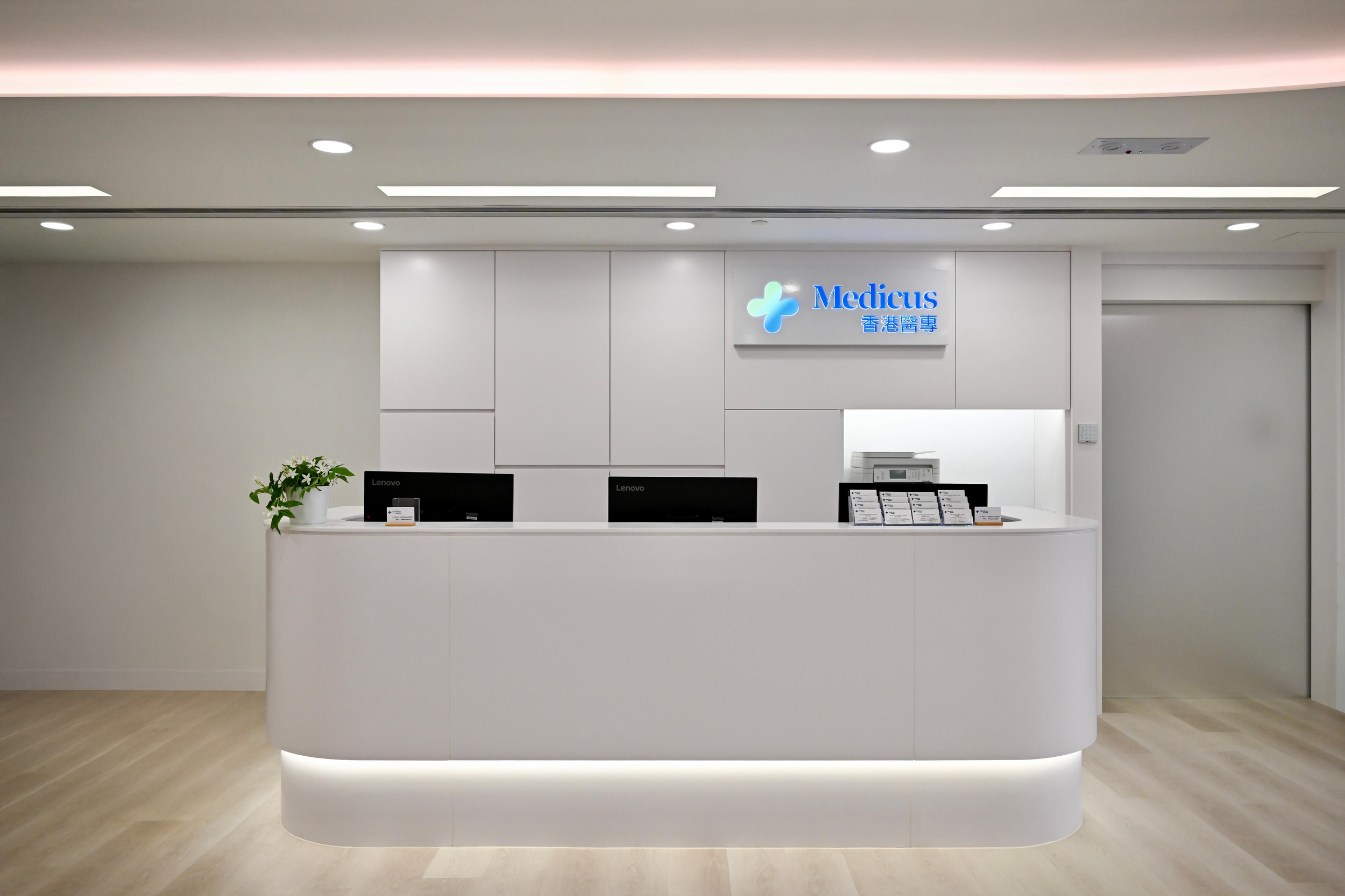 Modern hospital reception desk with a white curved counter, two black monitors, a potted plant, and a sign displaying 'Medicus' with green and blue logo, in a clean, minimalist design.