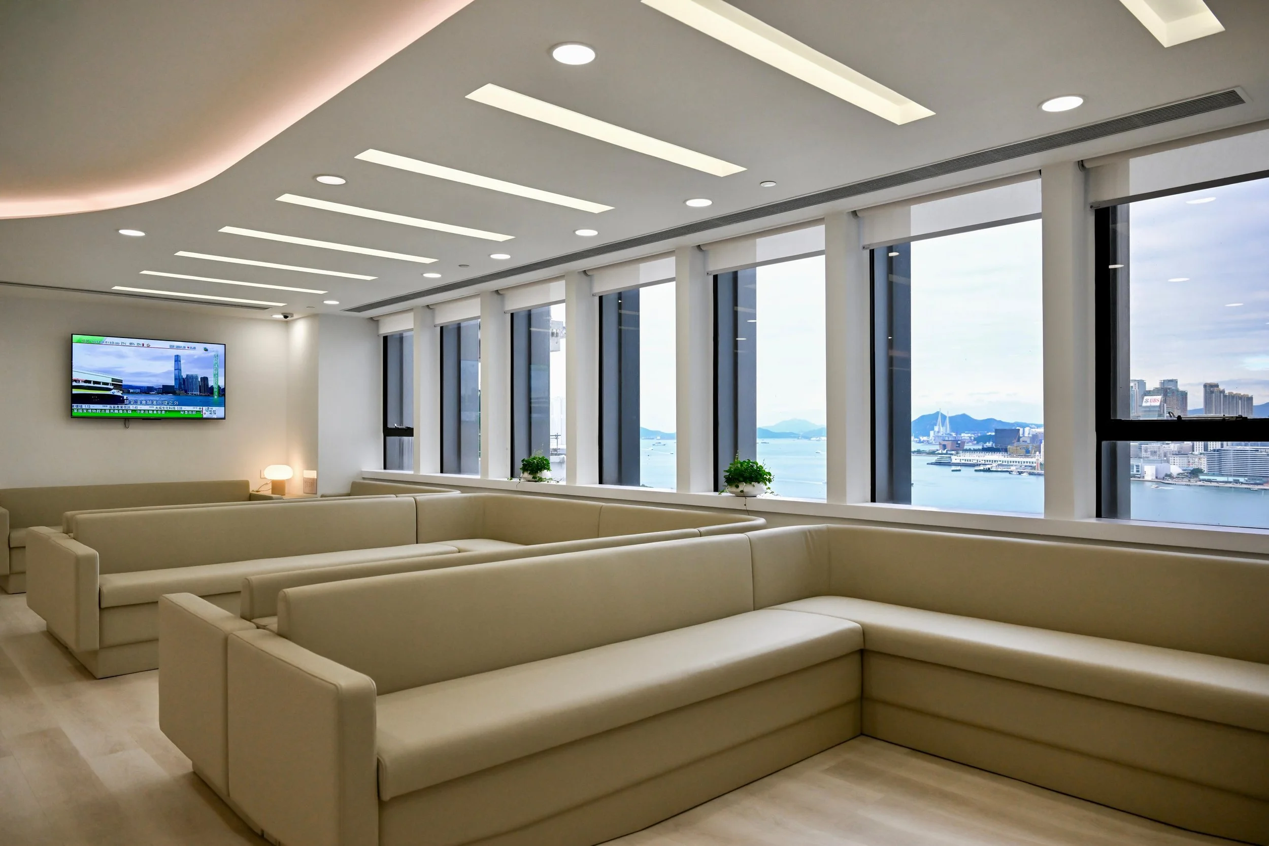 Modern lounge area with beige sofas, large windows showing a city skyline and body of water, and ceiling with recessed lighting.