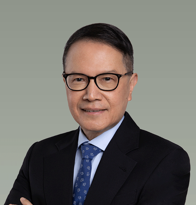 A professional headshot of a man with black hair and glasses, wearing a dark suit, light blue shirt, and patterned blue tie, against a muted green background.