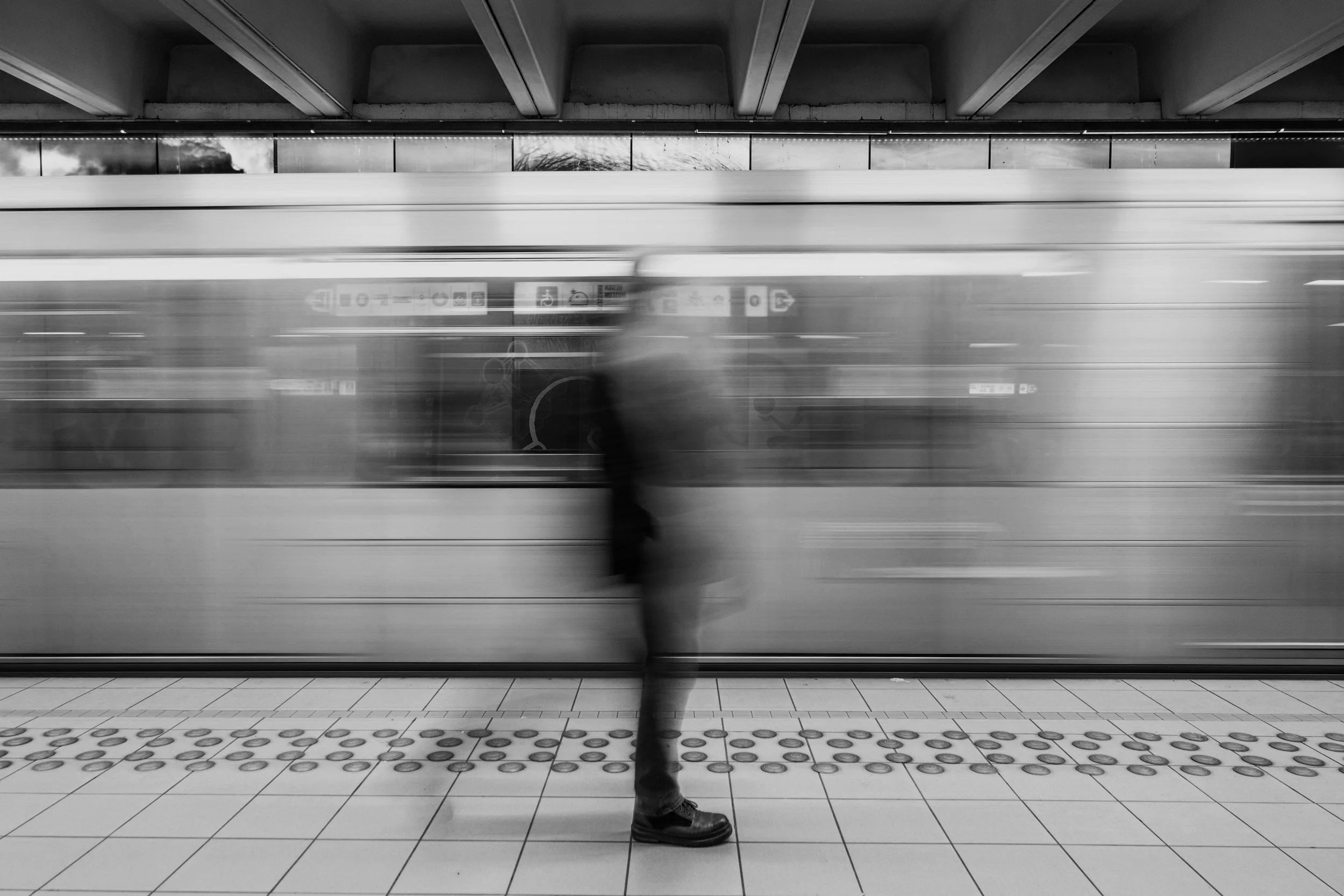 A person standing on a subway platform as a train speeds past, creating a blur effect in black and white.