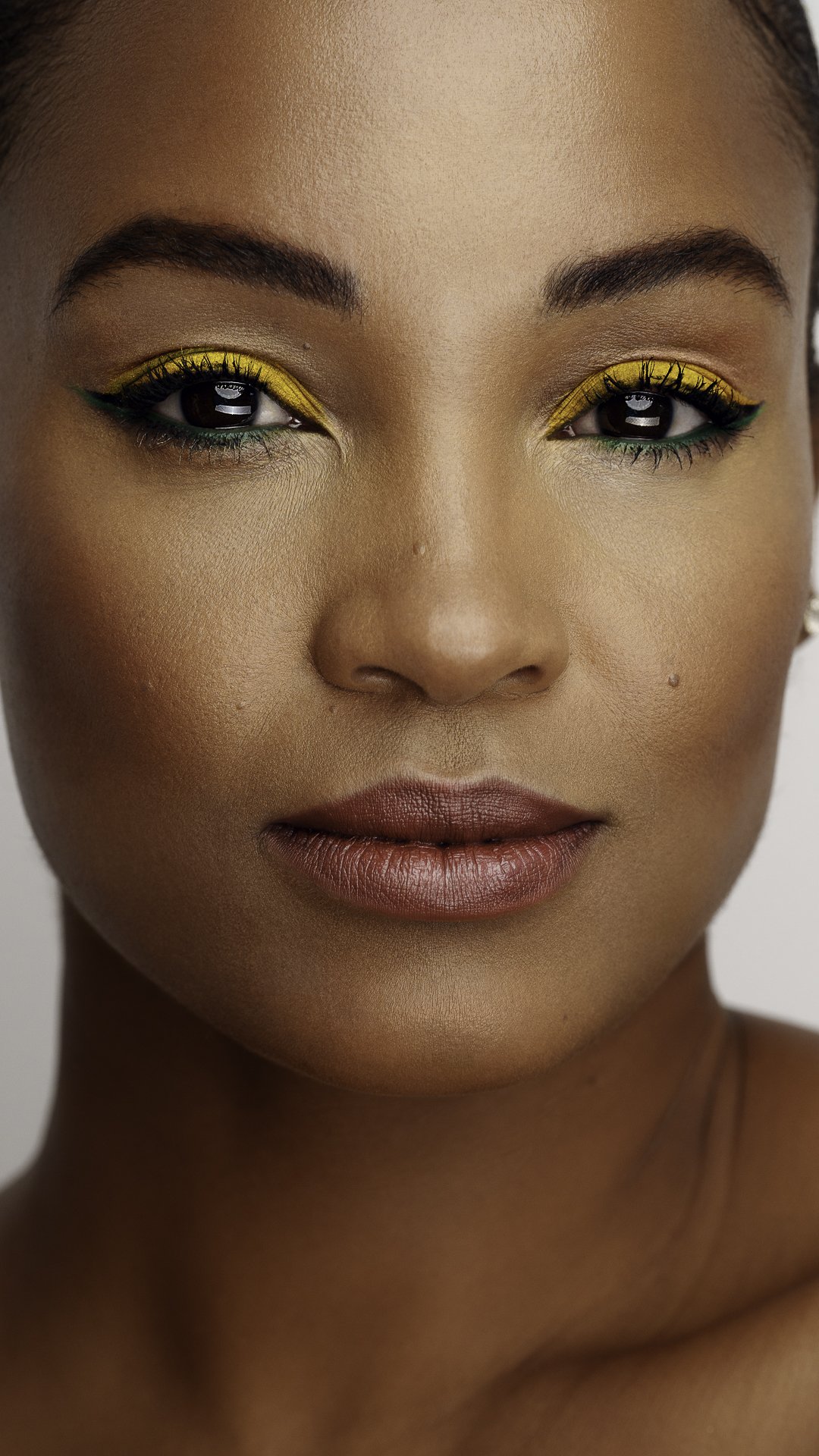 Close-up portrait of a woman with yellow and green eye makeup, well-defined eyebrows, and natural lips.