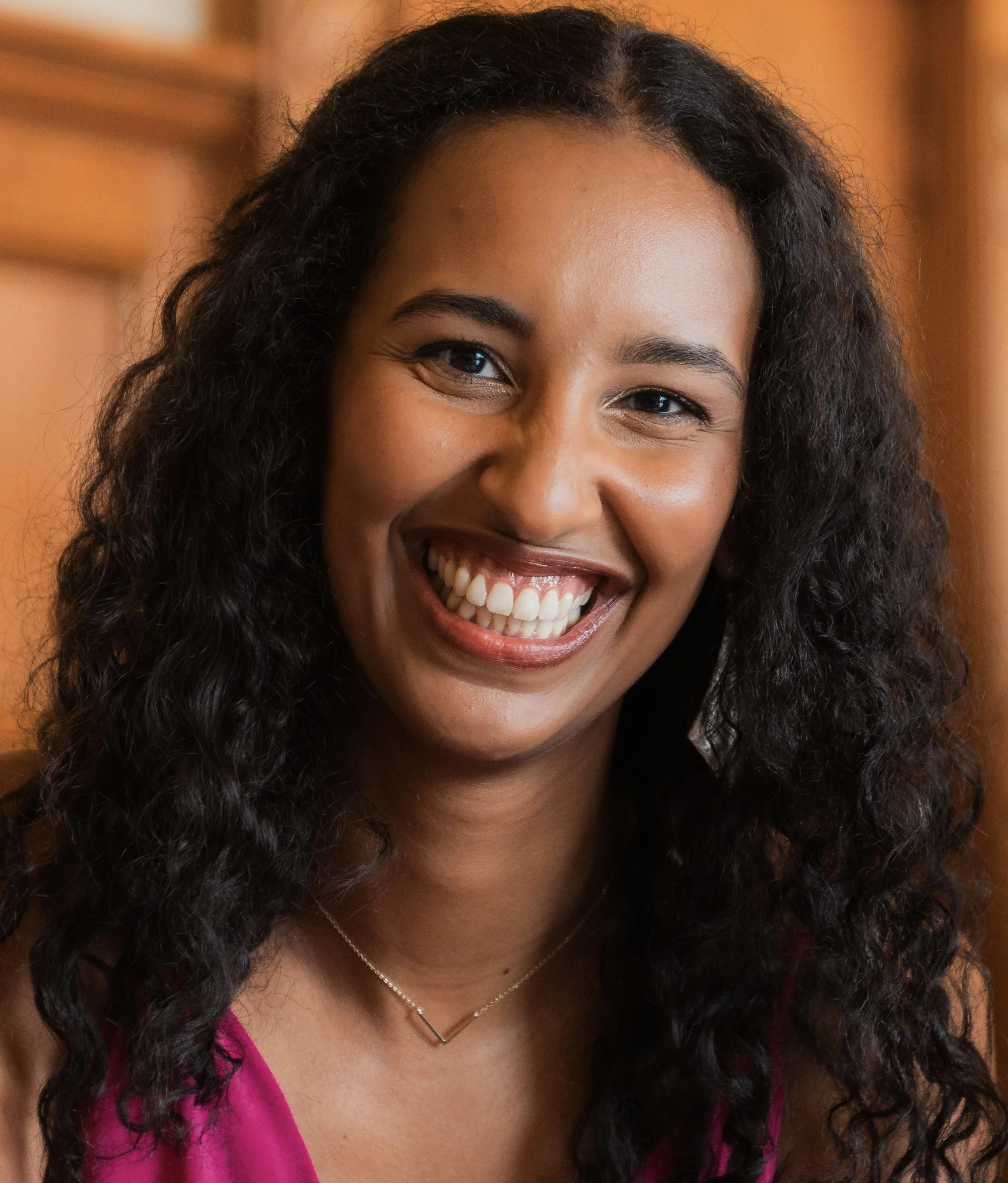 Close-up of a woman with curly black hair smiling warmly, wearing a pink top and a gold necklace, in front of a wooden background.