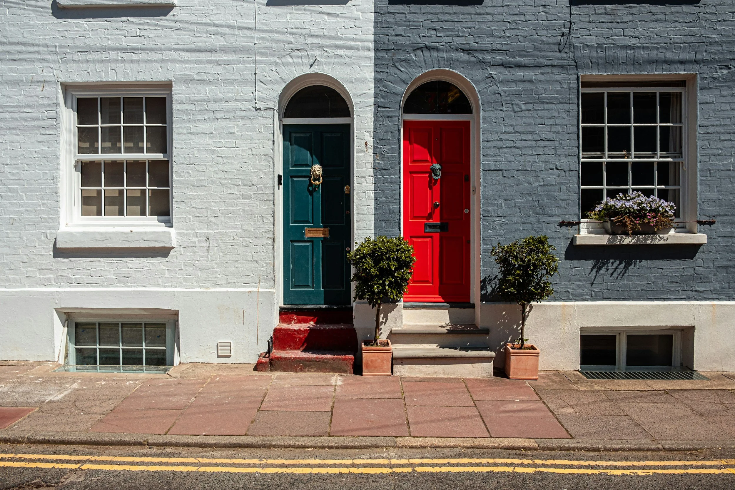 Two adjacent doors on a white brick building, with a green door on the left and a red door on the right, each decorated with a door knocker. There are potted bushes in front of each door and a window with a flower box on the right side. The building has steps leading up to both doors and a sidewalk in front.
