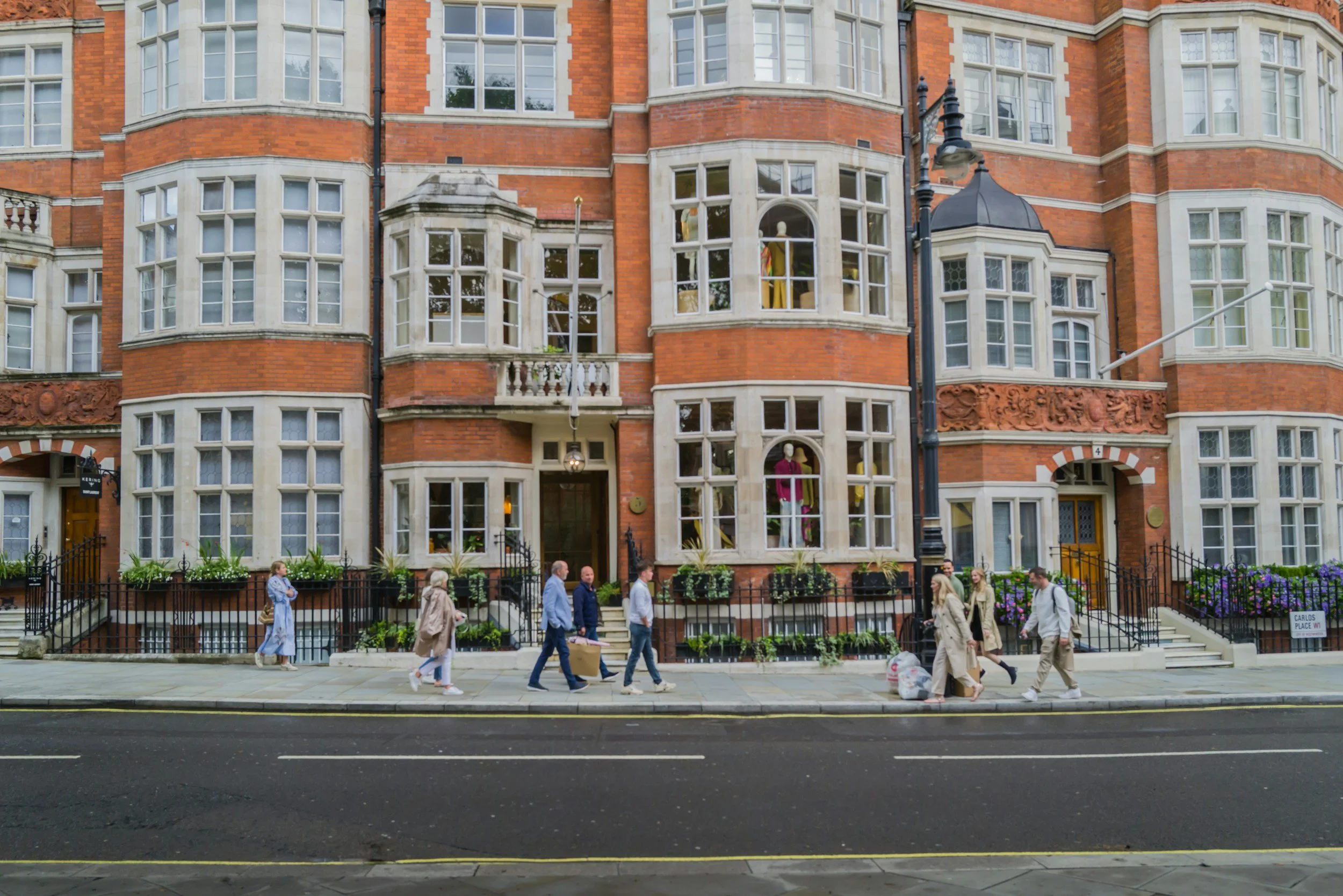People walking past a large red-brick apartment building with bay windows and ornate architectural details on a city sidewalk.