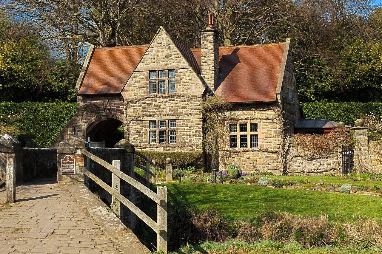 A historic stone house with a red tile roof, surrounded by greenery and a stone pathway leading to an arched entrance.