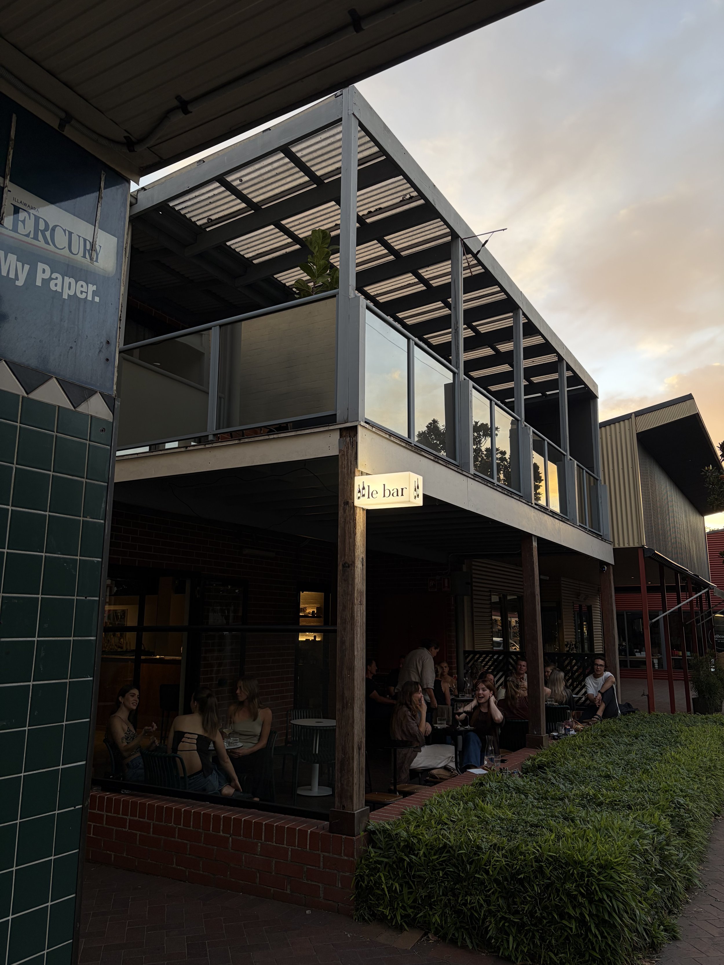 People sitting at outdoor tables of a restaurant or bar called 'ale bar' during sunset, with a two-story building featuring a balcony above.