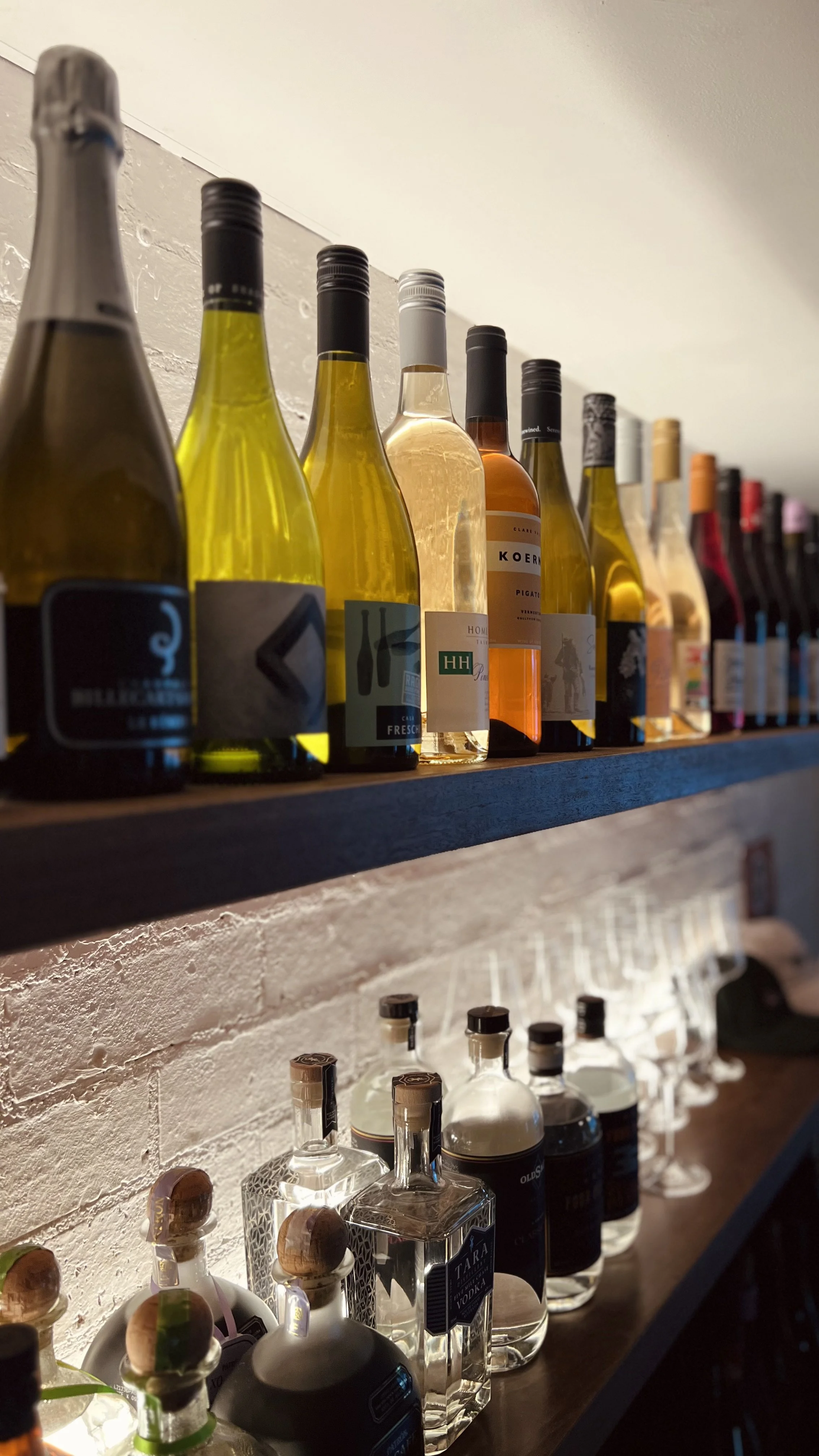 Collection of wine bottles on a wooden shelf with liquor bottles below against a white brick wall.