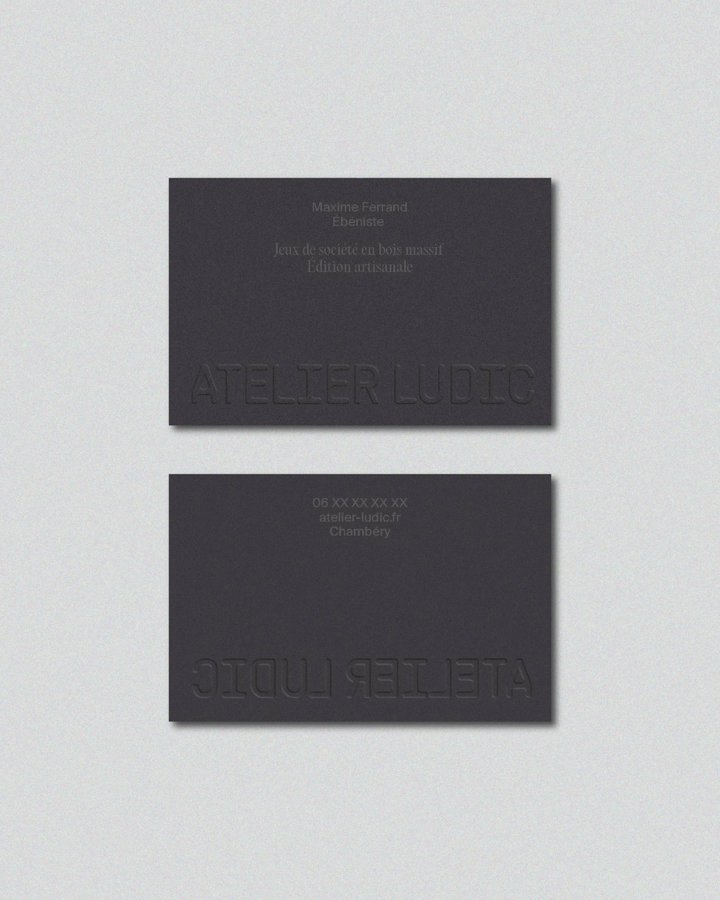 Deux cartes de visite noires avec du texte en relief, une située au-dessus de l'autre, sur un fond gris clair, probablement pour une entreprise ou un atelier artistique.