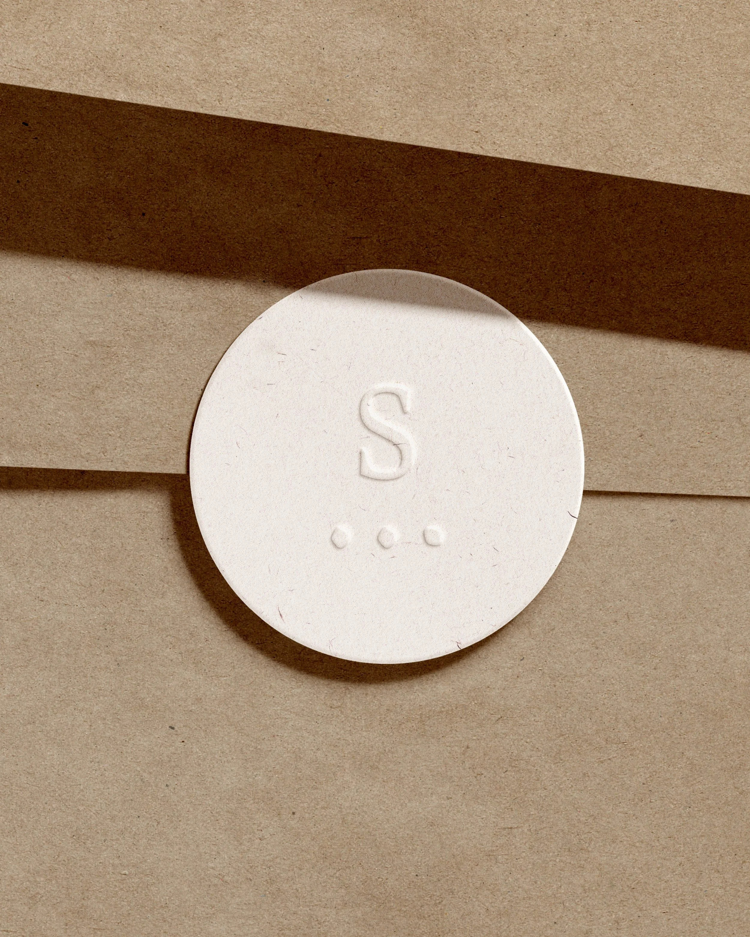 Une tablette ronde blanche avec le symbole 'S' et trois petits points en dessous, posée sur une surface beige avec des bandes de papier brun foncé en arrière-plan.