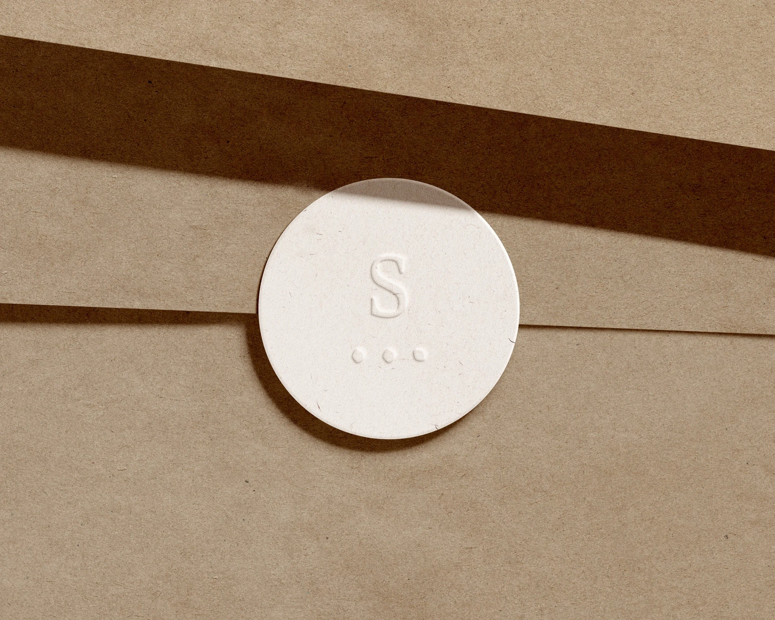 Disque blanc avec la lettre 'S' embossée, placé sur un fond de papier ou de carton beige.