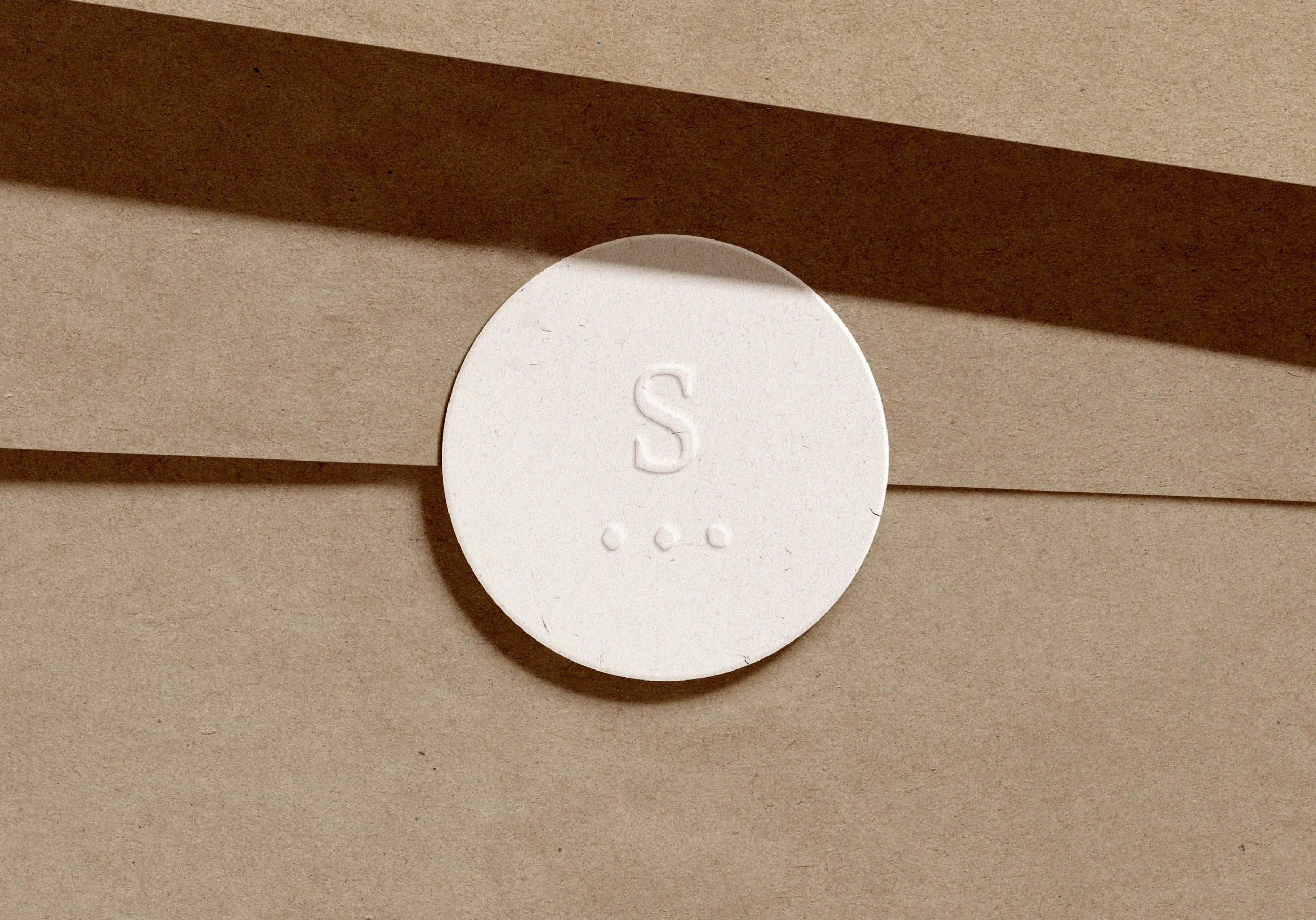 Un cachet rond blanc avec la lettre 'S' et trois points en dessous, posé sur une surface en papier marron.