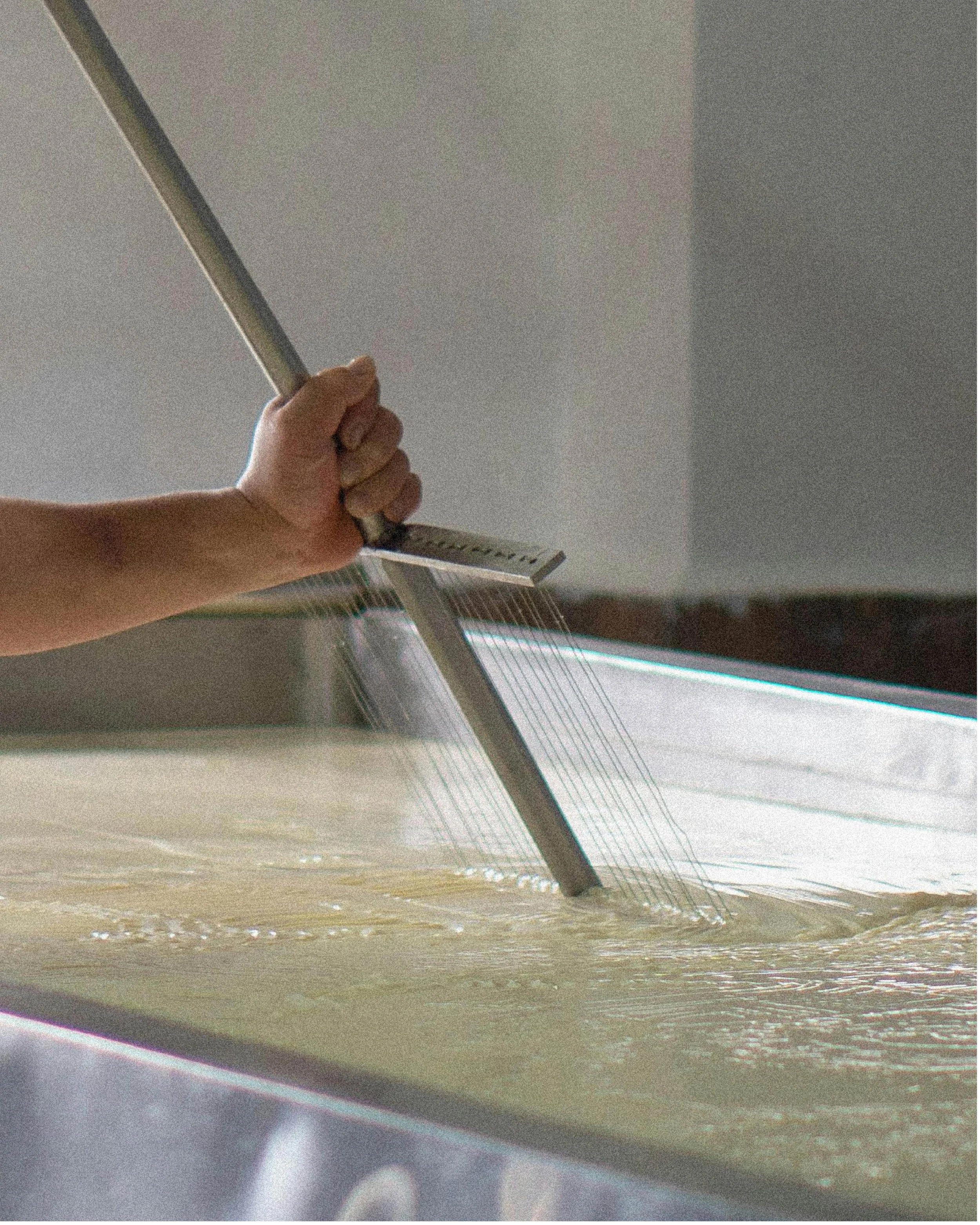 Une main manipulant un râteau pour étaler la pâte dans une grande cuve en acier inoxydable.