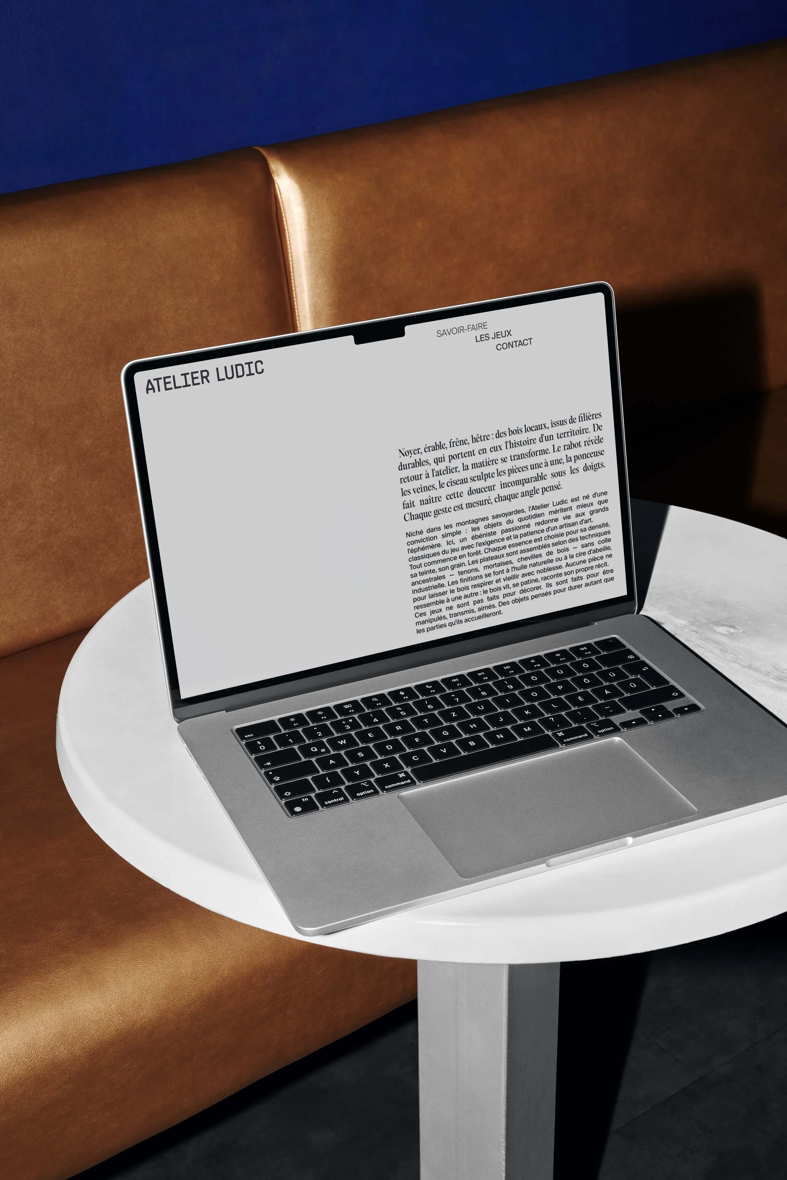Un ordinateur portable placé sur une petite table ronde blanche. L'écran affiche un texte en français, probablement une page web intitulée "Atelier Ludic". La table est devant un canapé en cuir brun et la pièce a un mur bleu en arrière-plan.