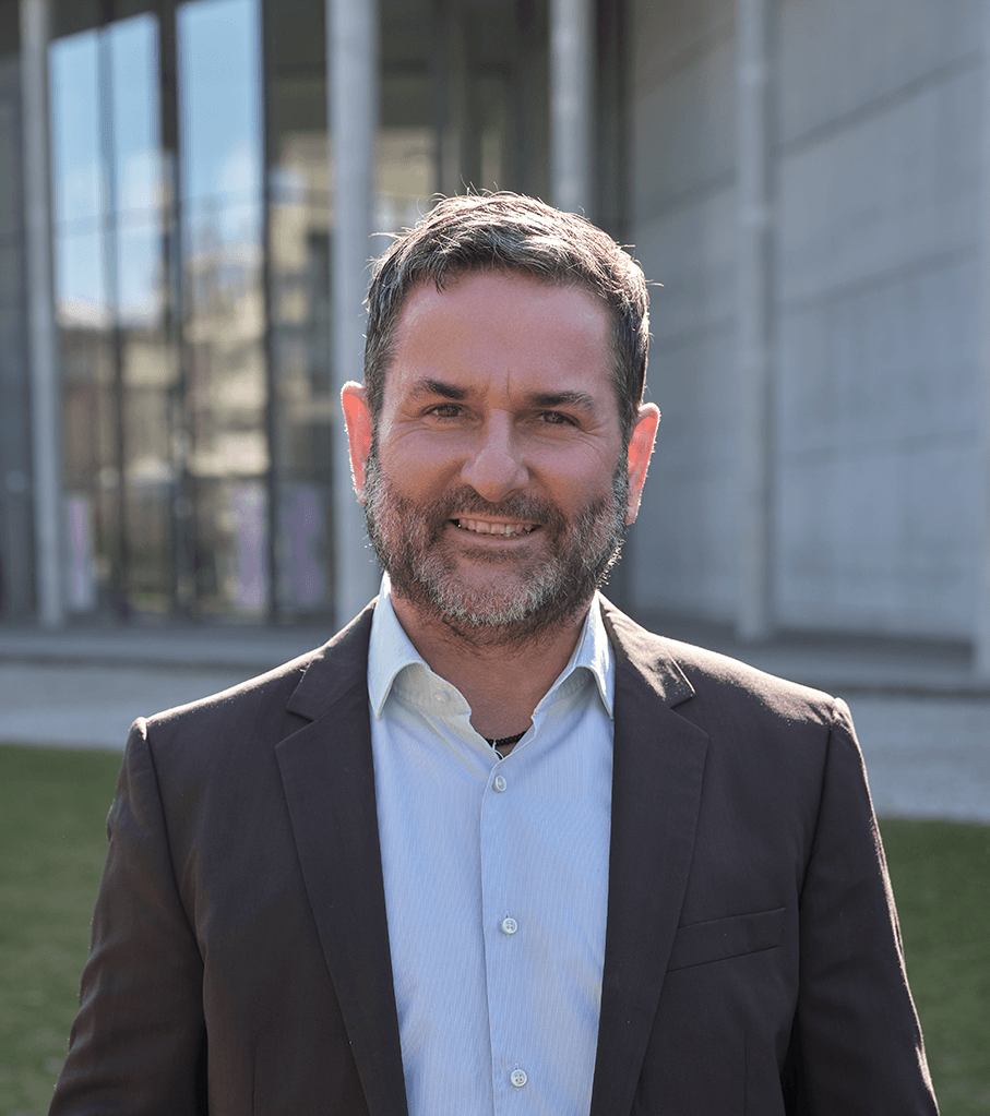 Mann mit blauem Hemd und braunem Sakko vor einem Gebäude, Sinnbild für mentale Gesundheit, Resilient und  Burnout-Prävention in München.png