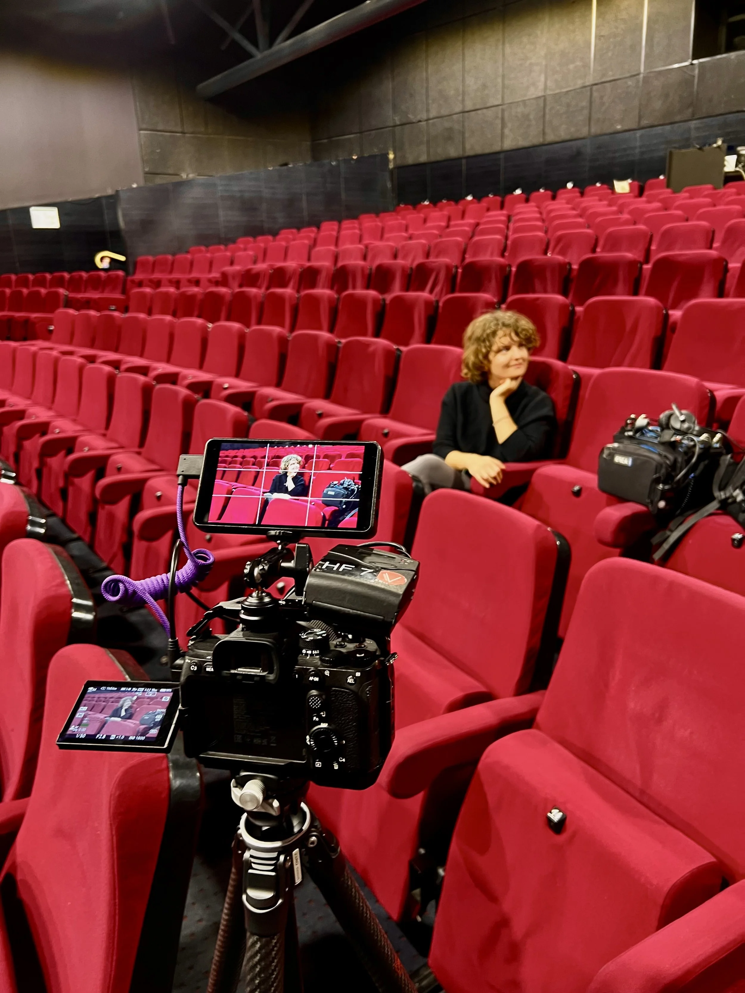 Salle de théâtre avec plusieurs rangées de sièges rouges, une femme assise parmi eux, appareil photo professionnel et caméra à l'avant pour la prise de vue.