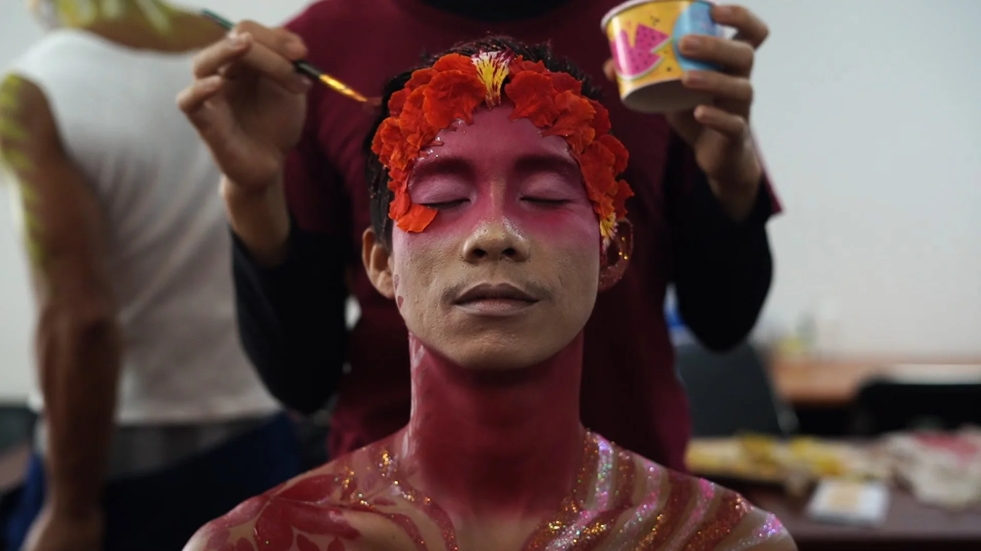 Une personne en train de faire du maquillage artistique, avec une peau entièrement peinte en rose et décorée de paillettes, portant un collier de fleurs rouges sur la tête.