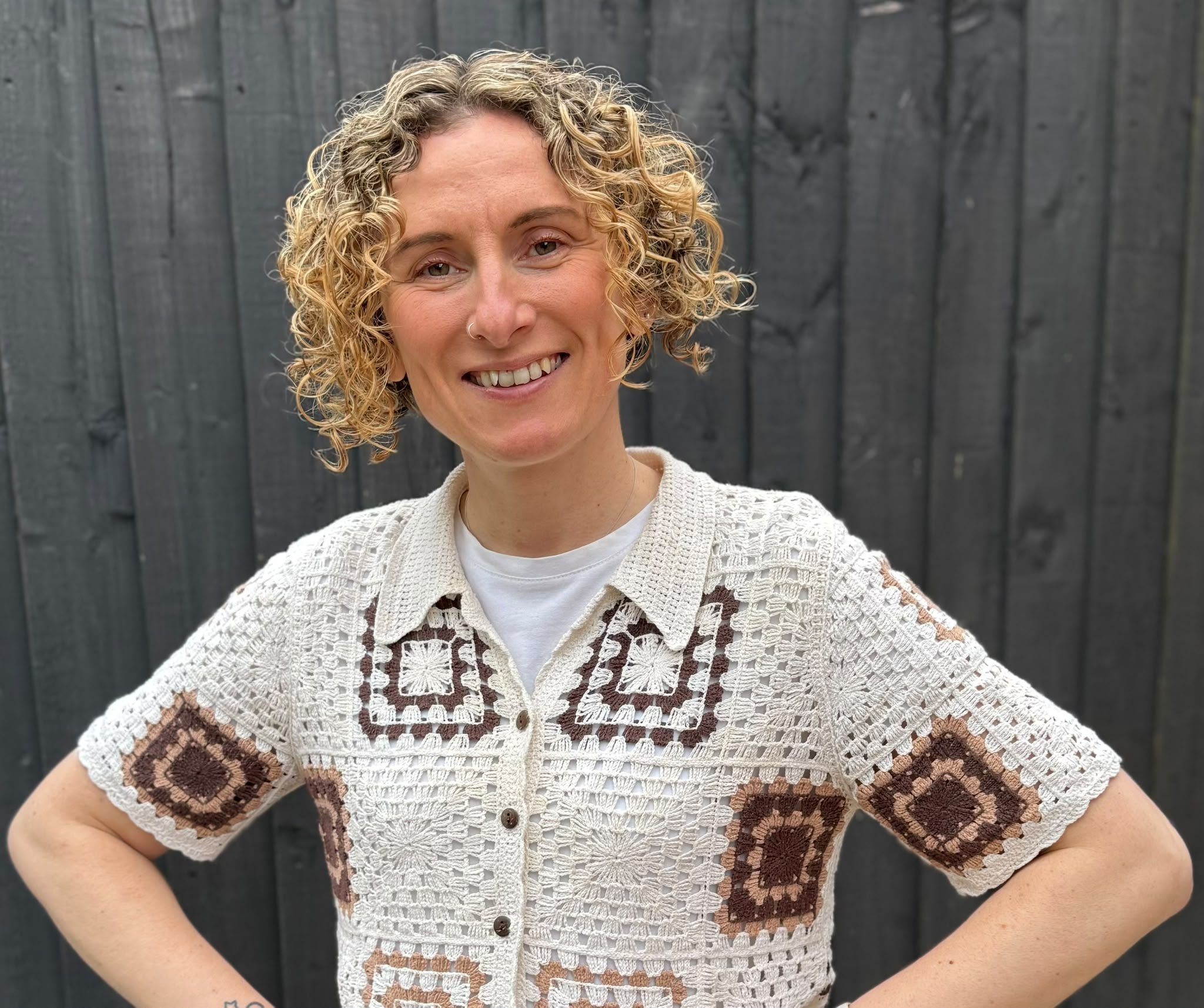 A woman with curly blonde hair is standing in front of a wooden fence and smiling