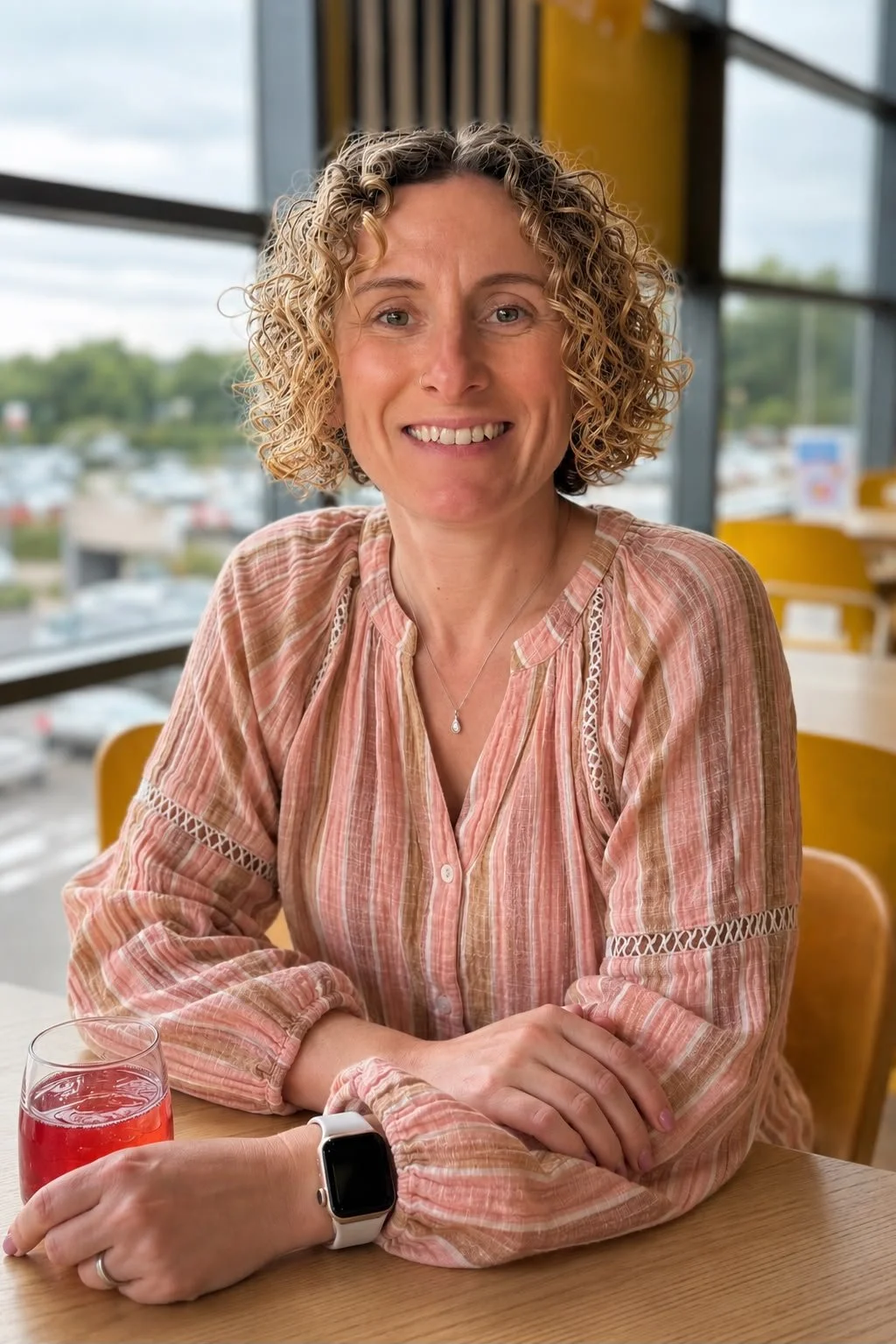 A woman with short curly blonde hair smiling, sitting at a table with a glass of pink beverage, wearing a pink and white striped blouse, inside a restaurant or cafe with large windows showing a parking lot and cloudy sky outside.