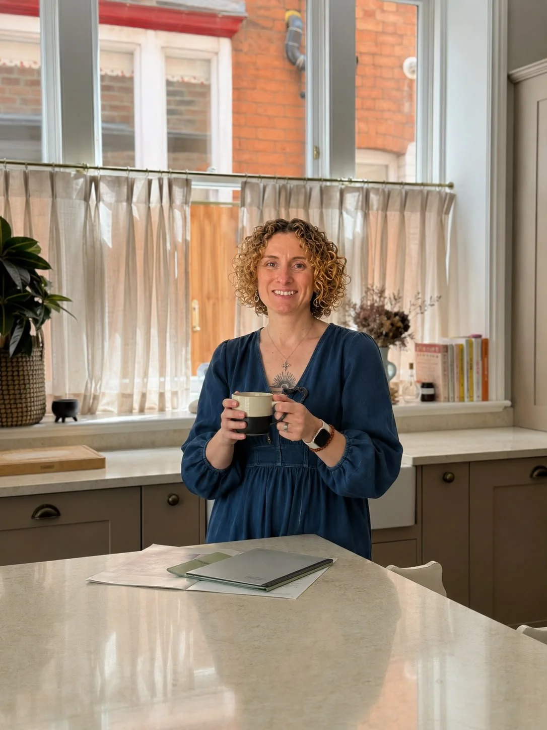 A woman with short curly hair is standing in a kitchen holding a mug and smiling