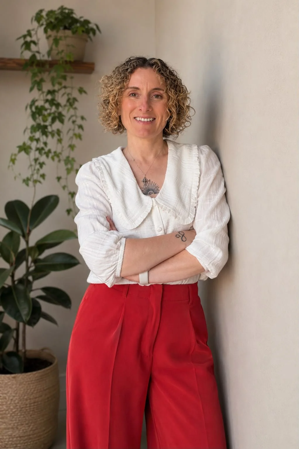 A woman with curly blonde hair, wearing a white blouse and red pants, standing indoors with her arms crossed, smiling, against a plain wall with a potted plant and a plant with trailing vines in the background.
