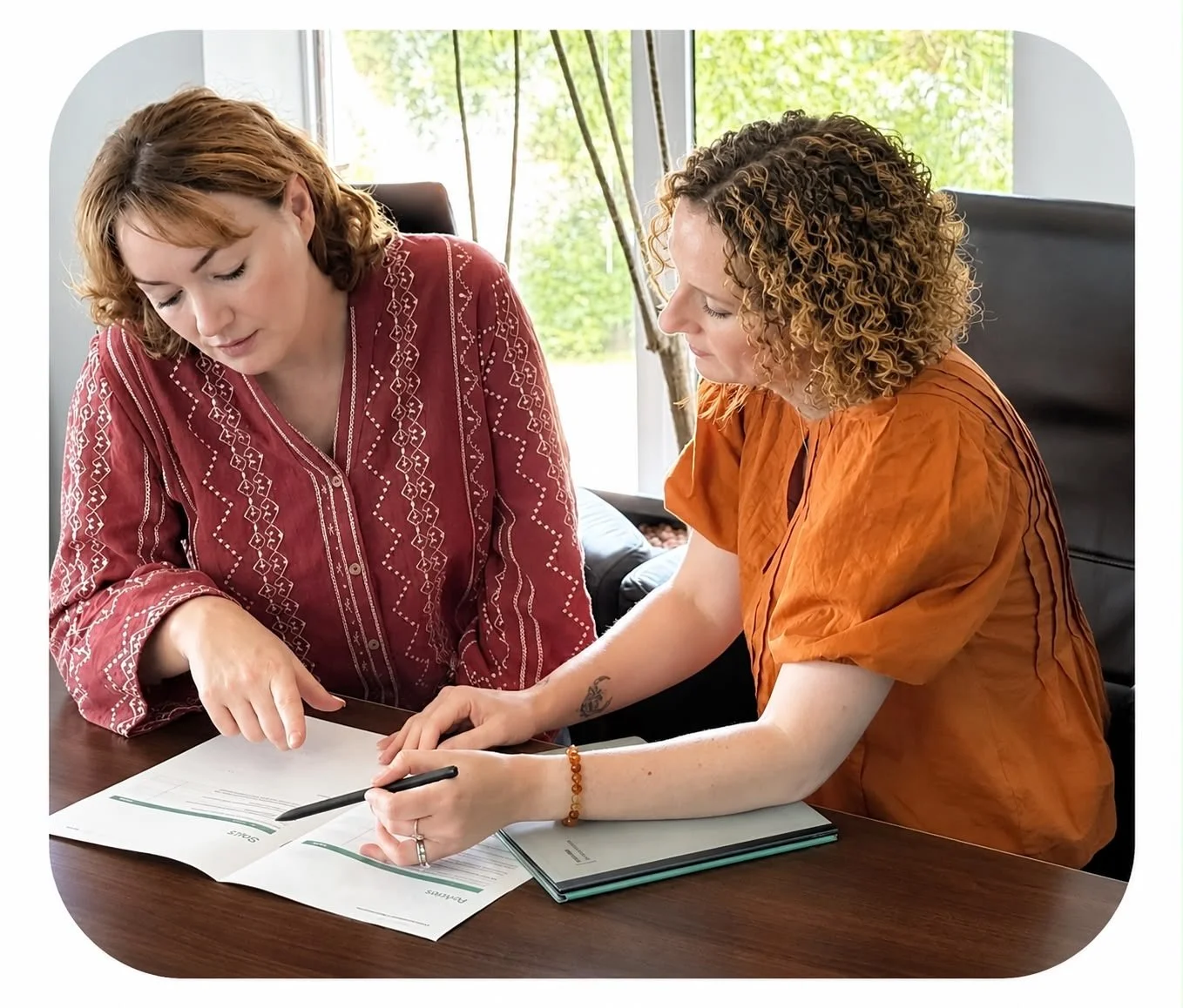 Two women sitting at a table, reviewing documents and using a pen together in a bright room with windows and greenery outside.
