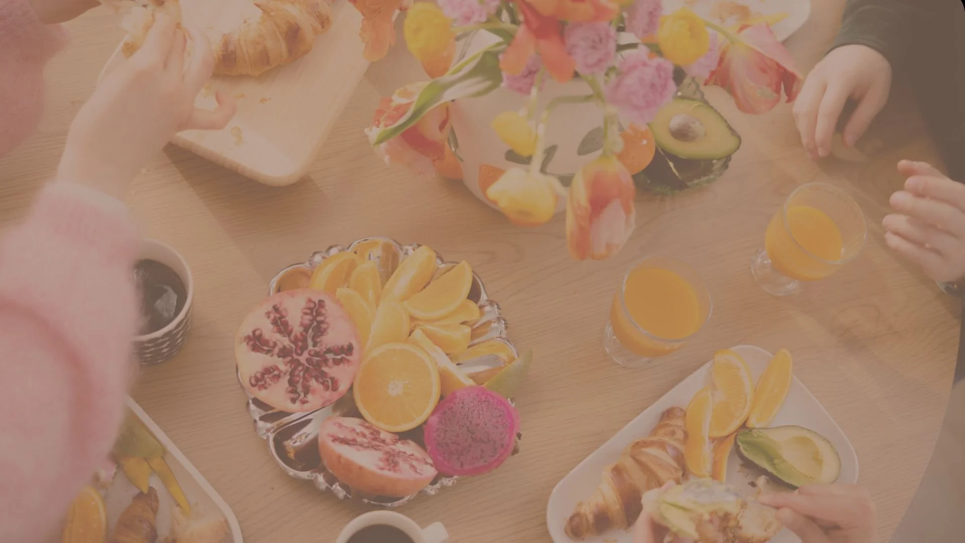 Pöytä, jonka päällä on erilaisia hedelmiä, kuten: appelsiineja, avokadoja ja croisantteja. Pöydässä on myös neljä lasillista appelsiinimehua ja kukkakimppu keskellä.