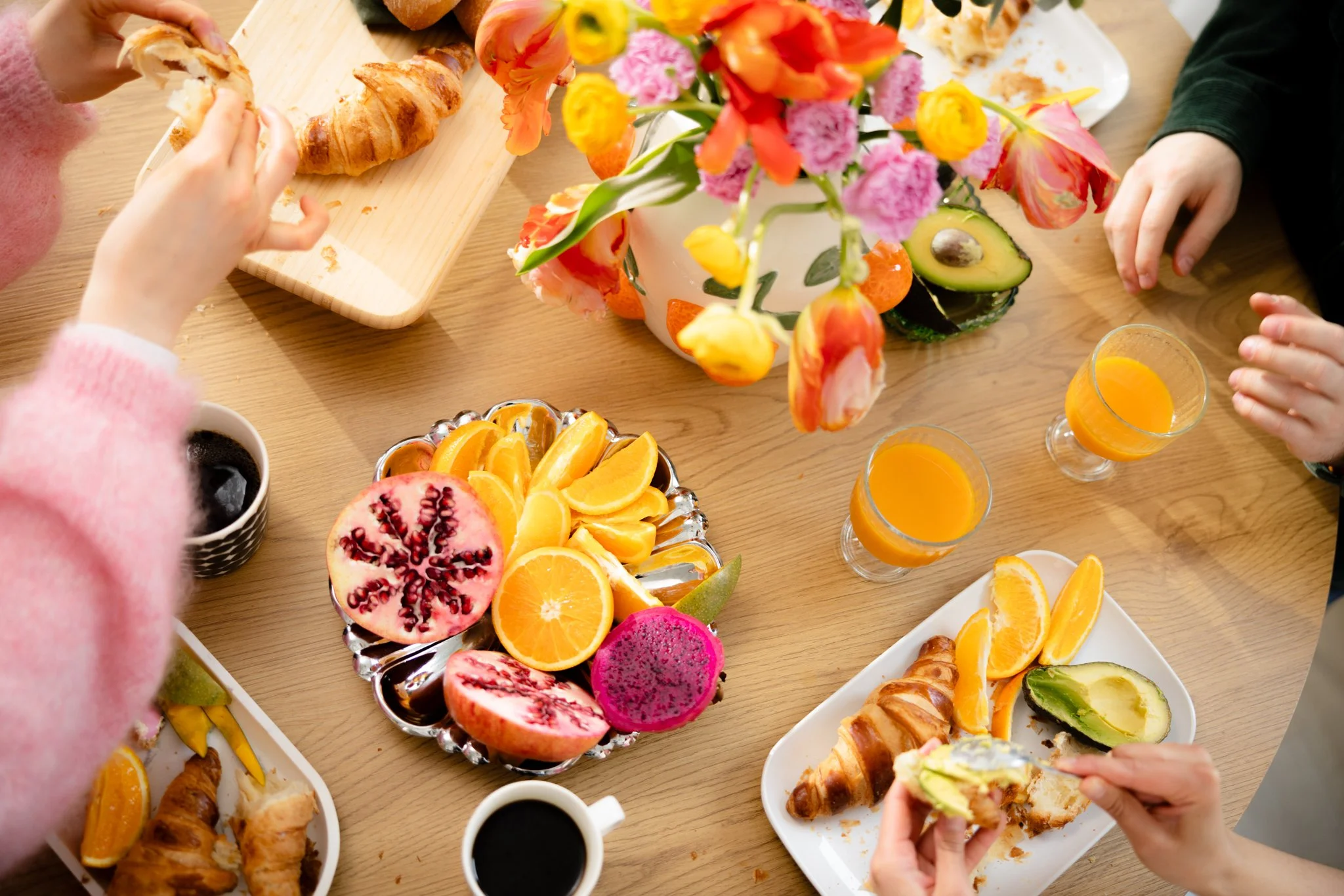 Pöytä, jolla on aamiaistarvikkeita, kuten croissanteja ja tuoreita hedelmiä. Appelsiineja, greippejä, avokadoja ja dragon-hedelmiä, lasillisia appelsiinimehua, kahvia sekä maljakko täynnä värikkäitä kukkia.