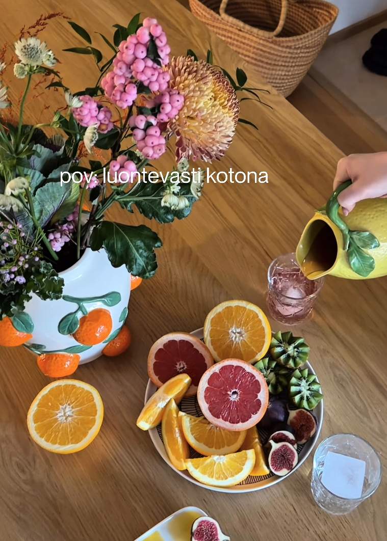 Pöytä, jolla on tuoreita hedelmiä, kuten appelsiineja, greippejä, viikunoita ja lasilliset mehua sekä maljakko täynnä värikkäitä kukkia.