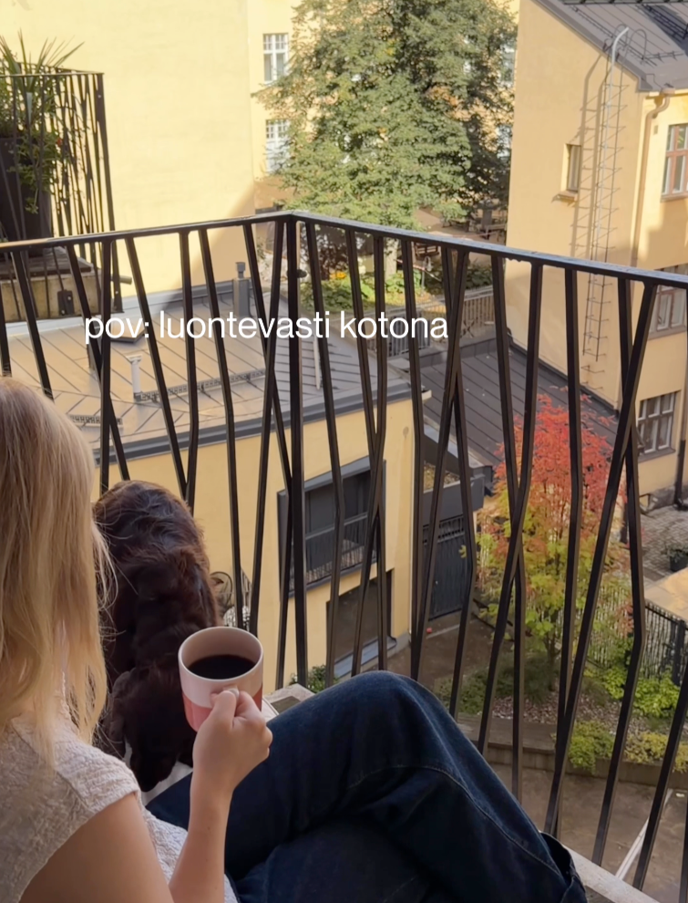 Nainen istuu parvekkeella ja juo kahvia, takana naapuritalojen rakennuksia ja puita syksyn värisinä.