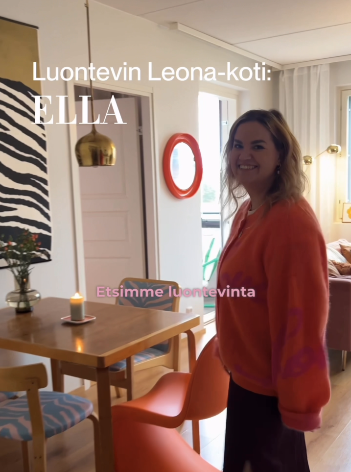 Nainen hymyilee sisätiloissa keittiössä. Takana pöytä, lamppu ja koriste-esineitä.