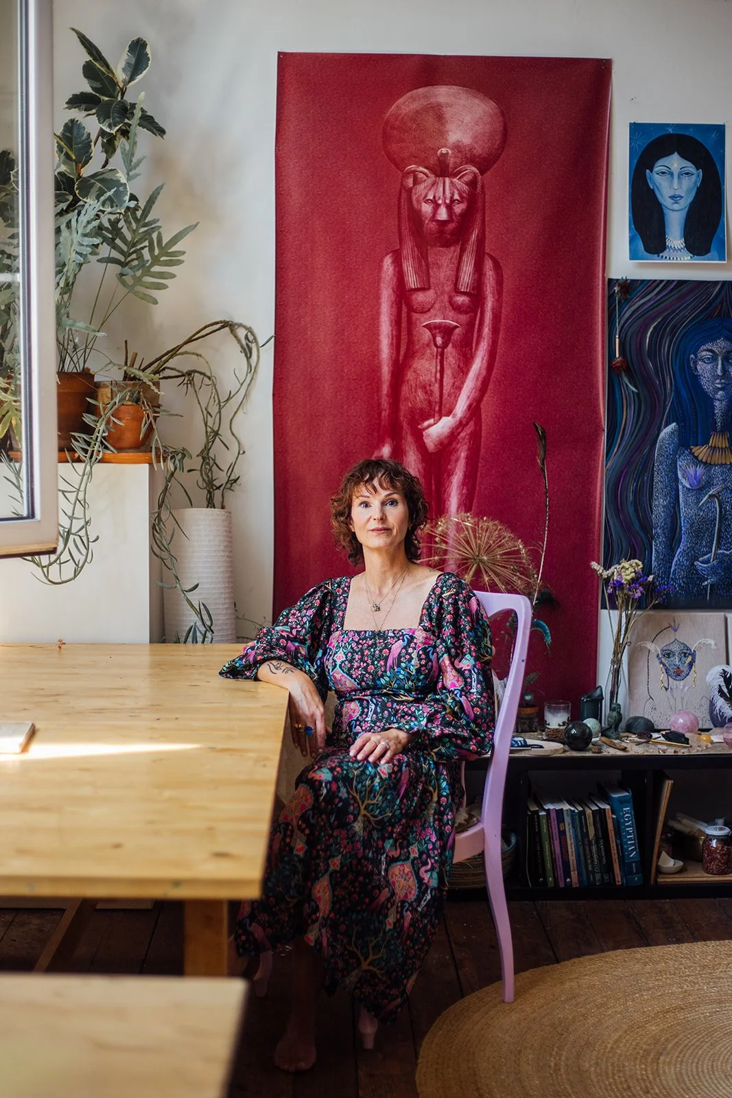 Een vrouw met korte, gekrulde haar zit aan een houten tafel in een artistiek ingerichte kamer met kleurrijke schilderijen en planten.
