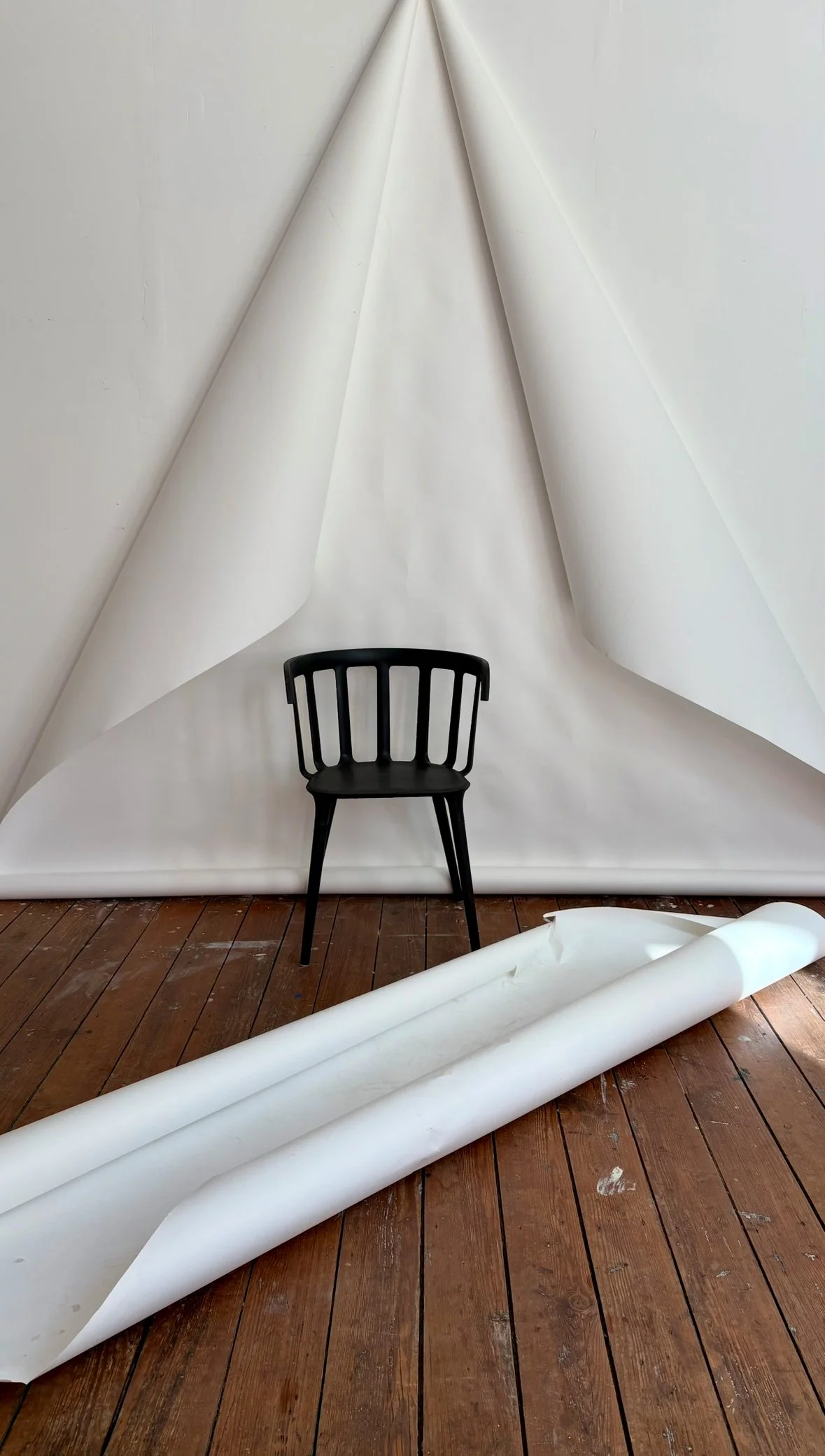 A black chair in front of a large white backdrop with rolled white paper on a wooden floor.