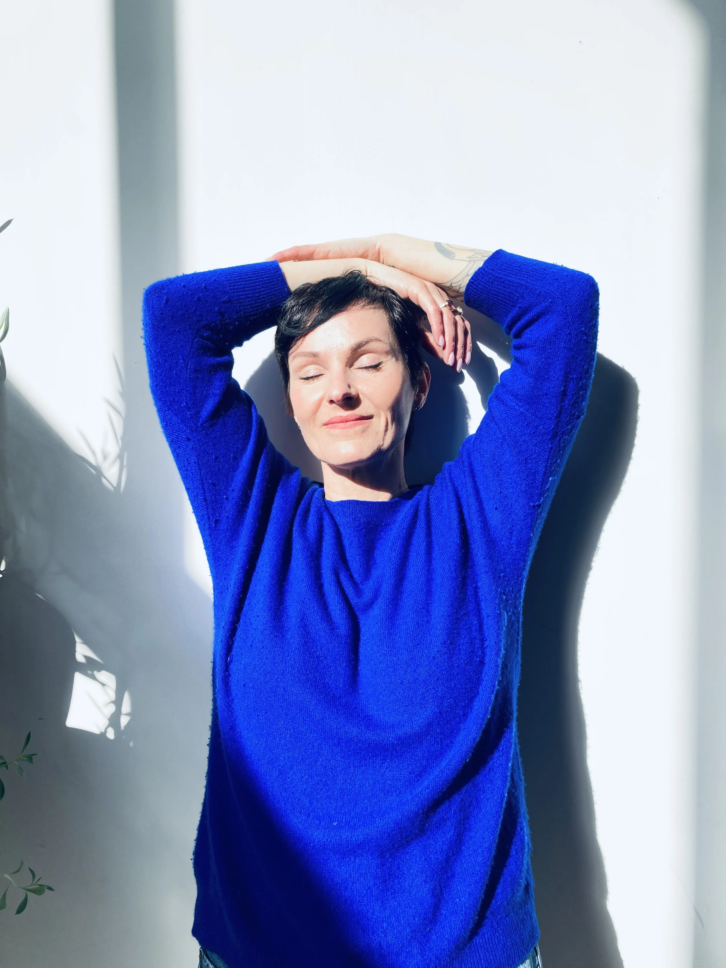 Person in blue sweater with eyes closed, arms raised above head, standing against a white wall with sunlight casting shadows.